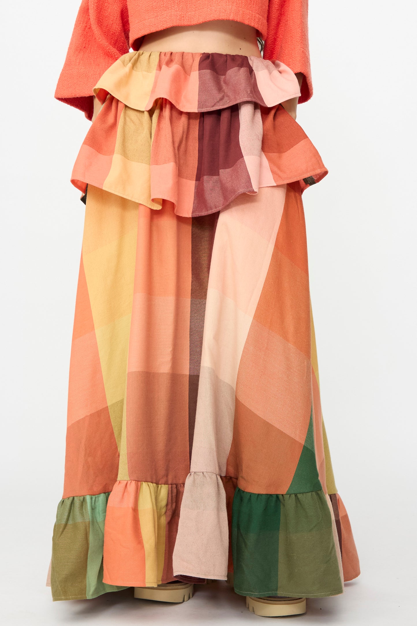 A person wearing Marrakshi Life’s The Tiered Ruffle Skirt in Plaid—a handmade maxi skirt from Morocco, featuring long, ruffled tiers and a vibrant geometric plaid pattern in orange, pink, green, and brown. Only the lower torso and legs are visible.