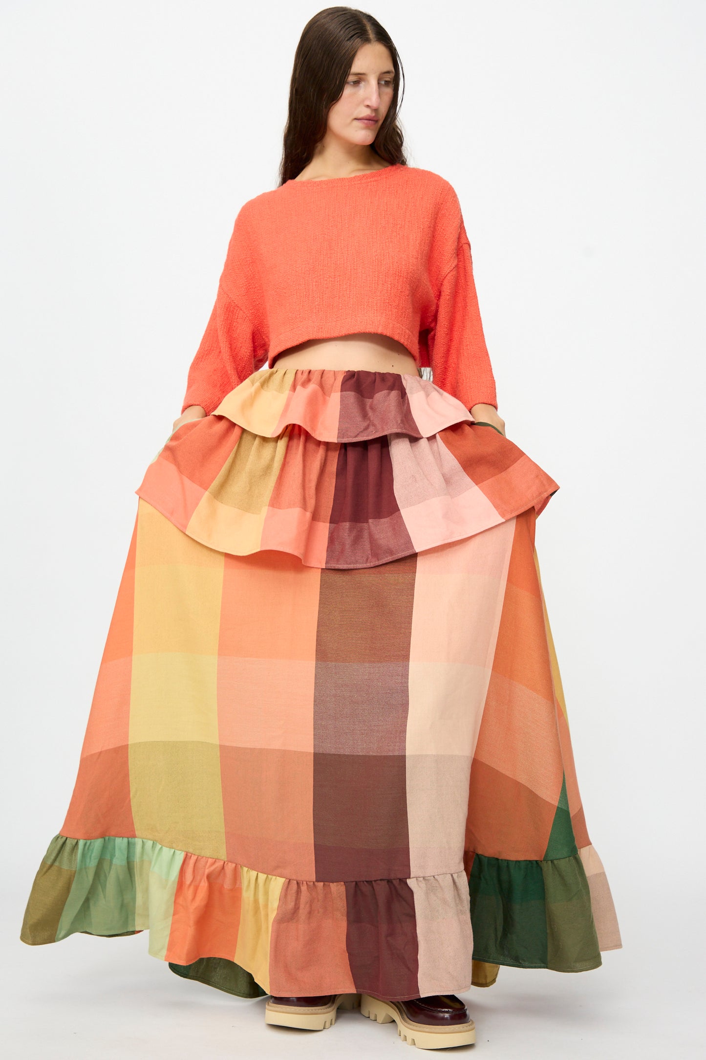 A woman wears Marrakshi Life’s The Tiered Ruffle Skirt in Plaid—a long, handwoven cotton maxi skirt with ruffle details, handmade in Morocco—paired with an orange cropped sweater, standing against a plain white background.