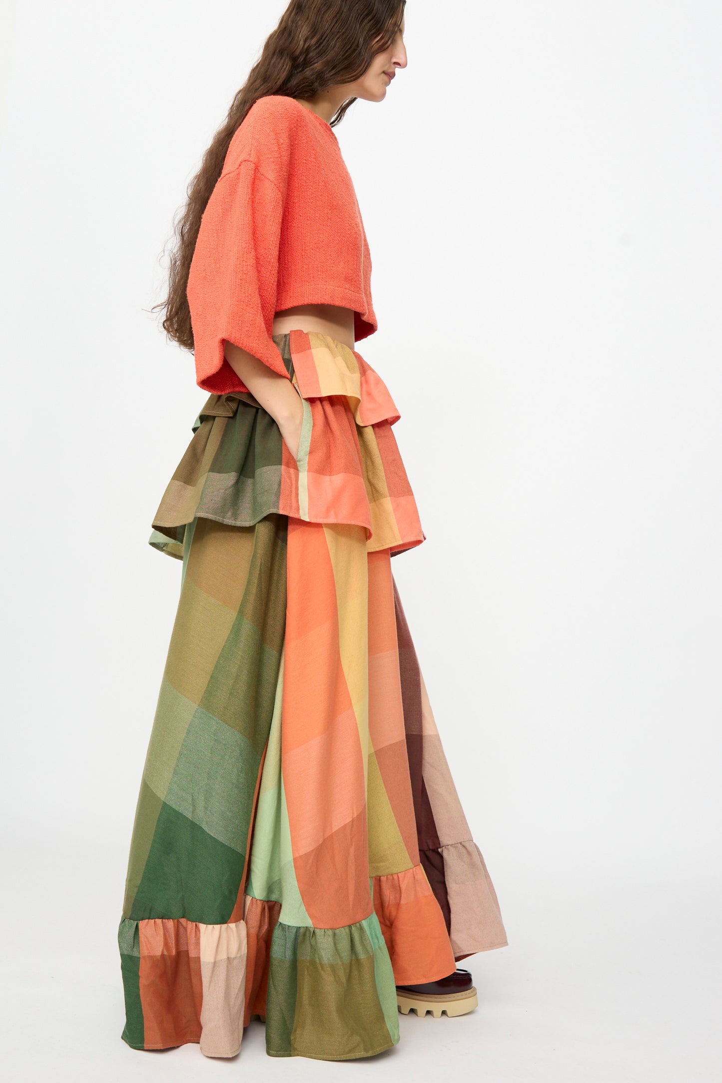 A person wearing The Tiered Ruffle Skirt in Plaid by Marrakshi Life, a wide-leg, multi-colored patchwork skirt with ruffle details, handmade in Morocco, stands against a plain white background.