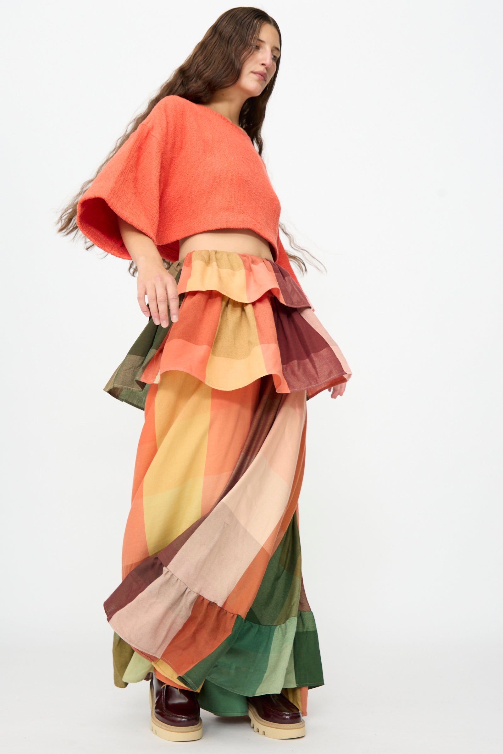 A person wearing a loose coral crop top and Marrakshi Life’s The Tiered Ruffle Skirt in Plaid, paired with platform shoes, stands against a plain white background.