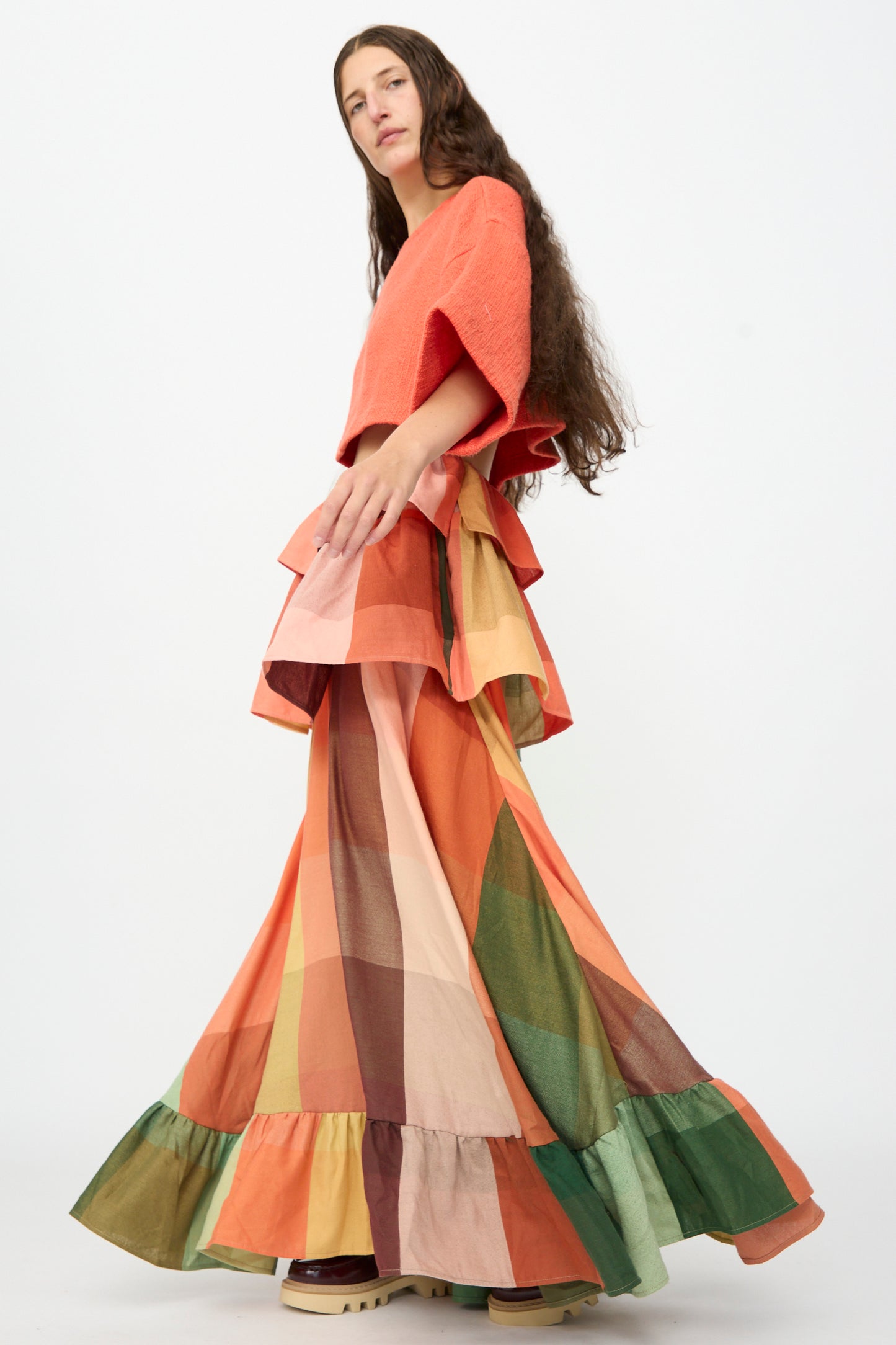 A person with long brown hair wears an orange top and The Tiered Ruffle Skirt in Plaid, a long, colorful handwoven cotton maxi skirt with ruffle details by Marrakshi Life, handmade in Morocco, standing against a plain white background.