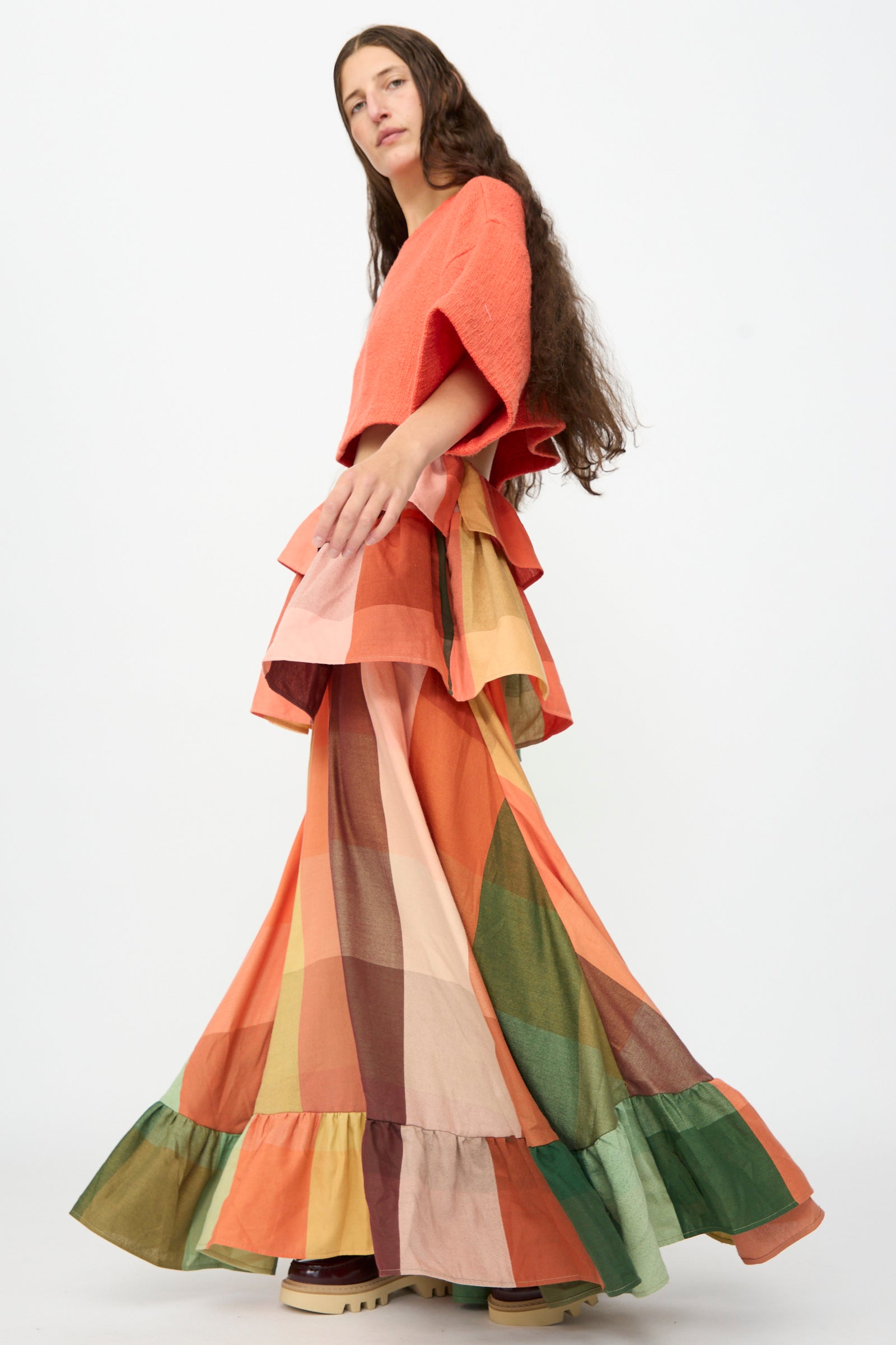 A person with long brown hair wears an orange top and The Tiered Ruffle Skirt in Plaid, a long, colorful handwoven cotton maxi skirt with ruffle details by Marrakshi Life, handmade in Morocco, standing against a plain white background.