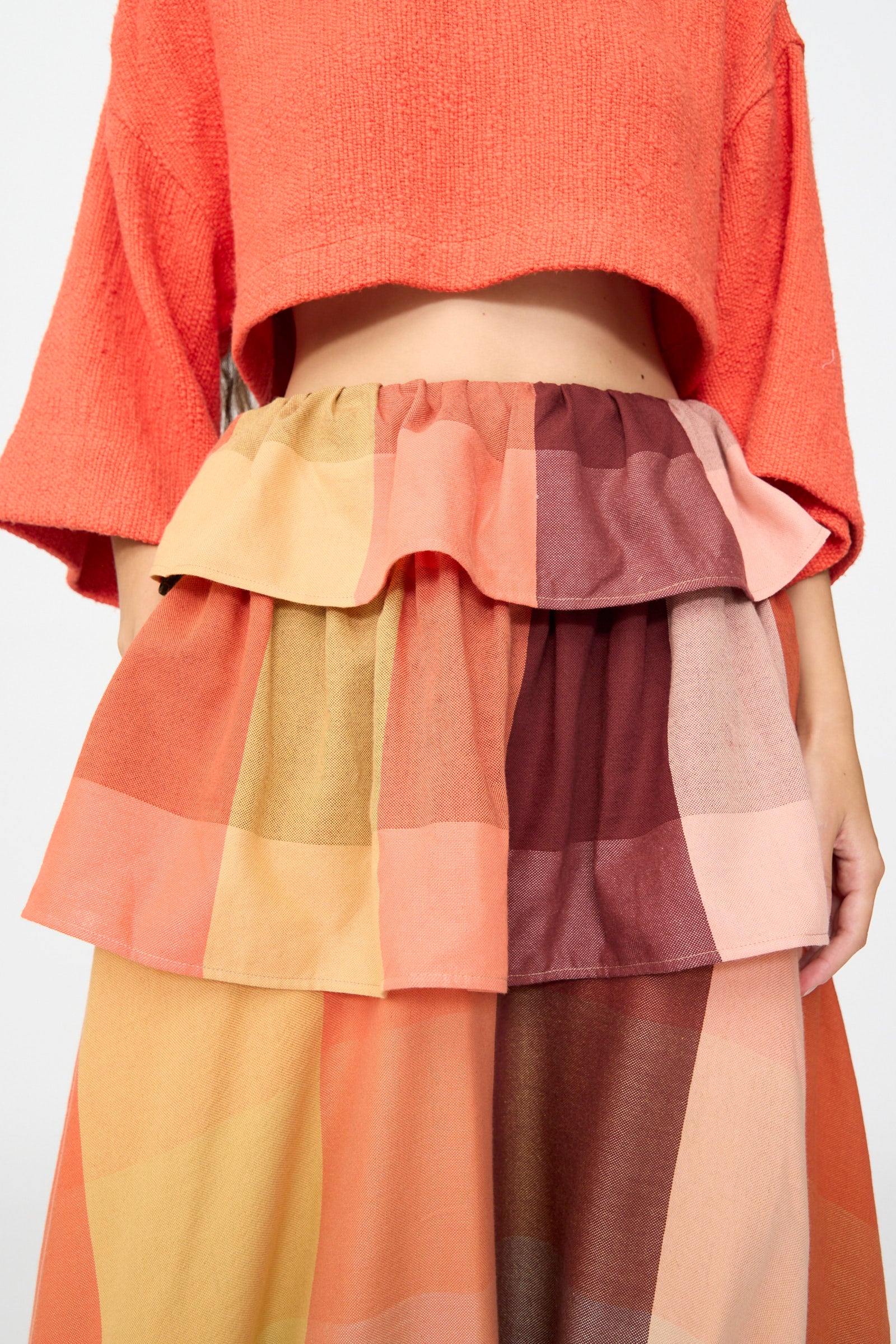 Wearing a cropped, textured coral top, the person pairs it with Marrakshi Life’s The Tiered Ruffle Skirt in Plaid—a high-waisted, handwoven cotton maxi skirt featuring large multicolored squares in warm tones, handmade in Morocco.
