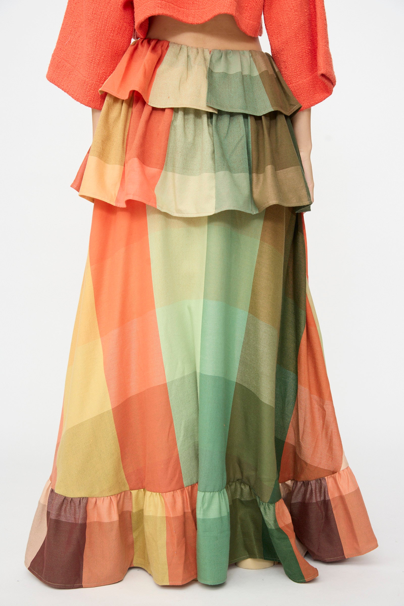 Wearing Marrakshi Life’s The Tiered Ruffle Skirt in Plaid—a colorful, handwoven cotton maxi skirt crafted in Morocco with layered ruffles and bold plaid patterns in green, orange, yellow, and brown—paired with a matching orange top.