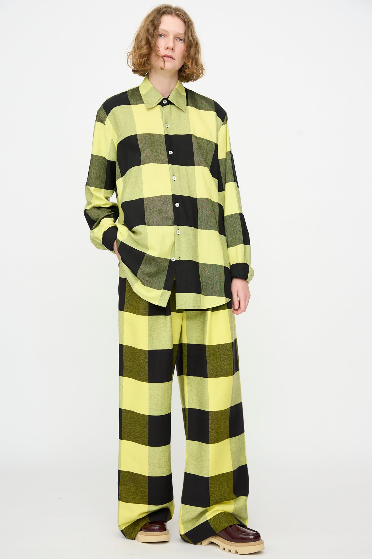 A person with shoulder-length wavy hair wears The Trouser in Plaid Black and Sunny by Marrakshi Life, styled with a matching long-sleeved button-up shirt in a bold black and neon yellow buffalo check pattern, set against a white background.
