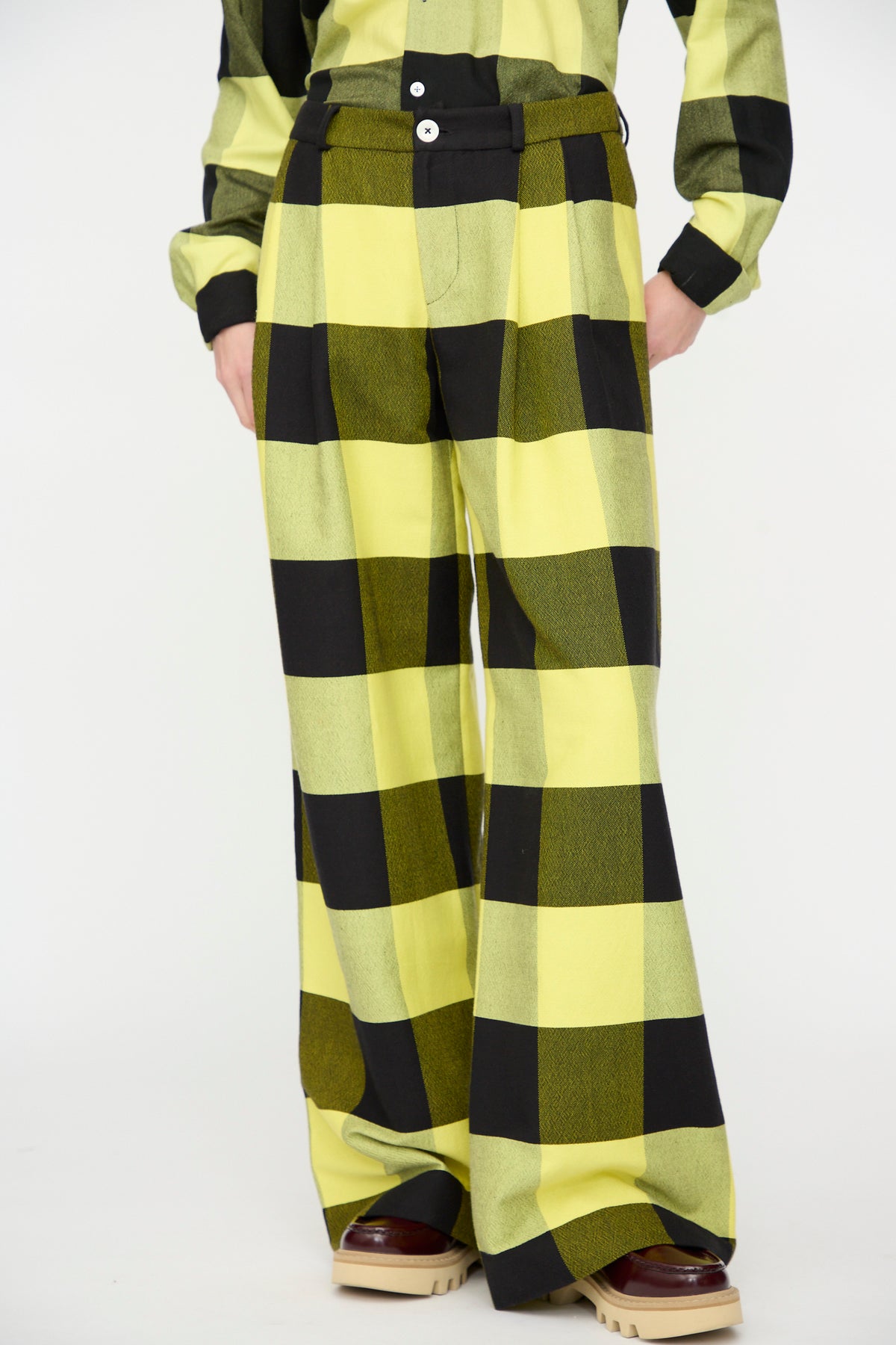 A person stands against a plain white background wearing Marrakshi Life’s The Trouser in Plaid Black and Sunny, featuring a wide-leg cotton silhouette, paired with a matching long-sleeve shirt and platform shoes.