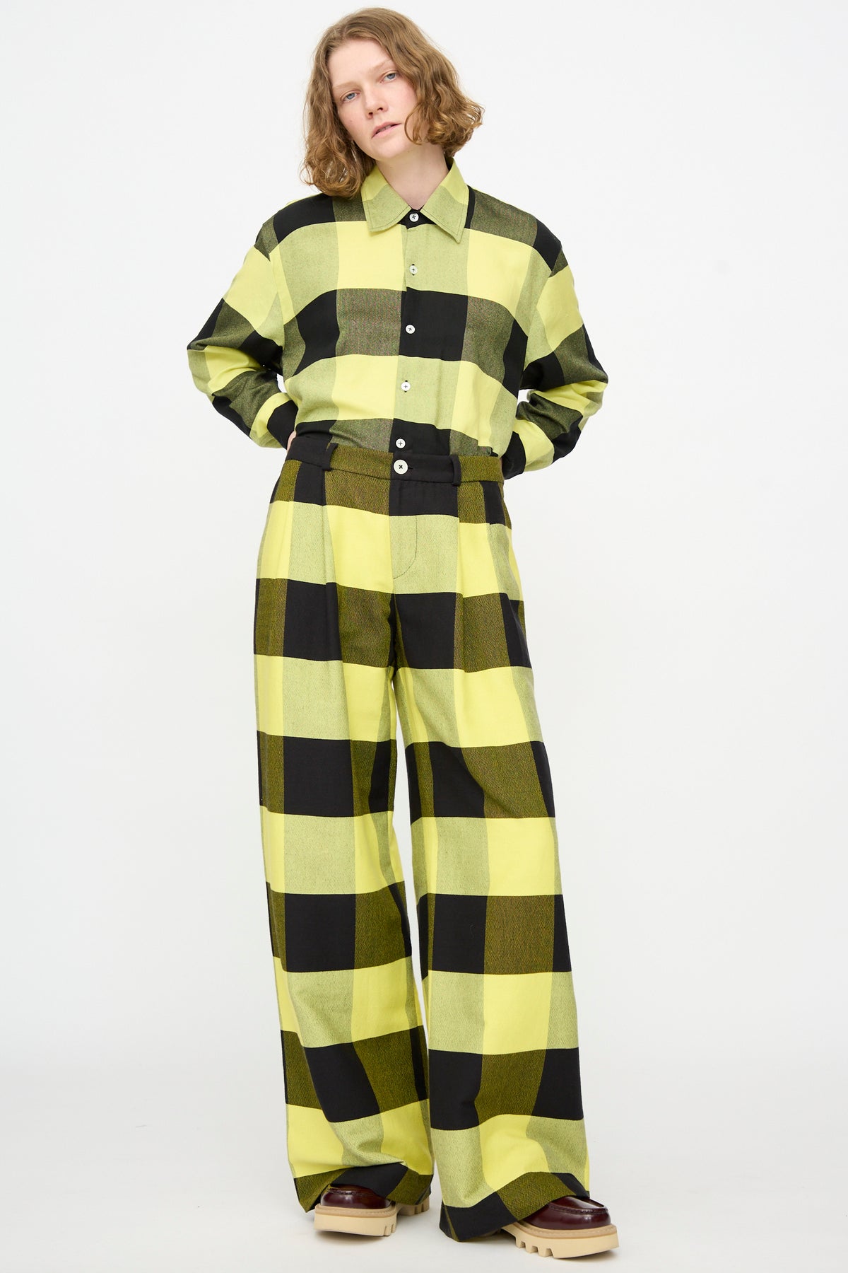 A person stands against a white background wearing Marrakshi Life’s The Trouser in Plaid Black and Sunny, styled with a matching yellow and black checkered button-up shirt and brown shoes.