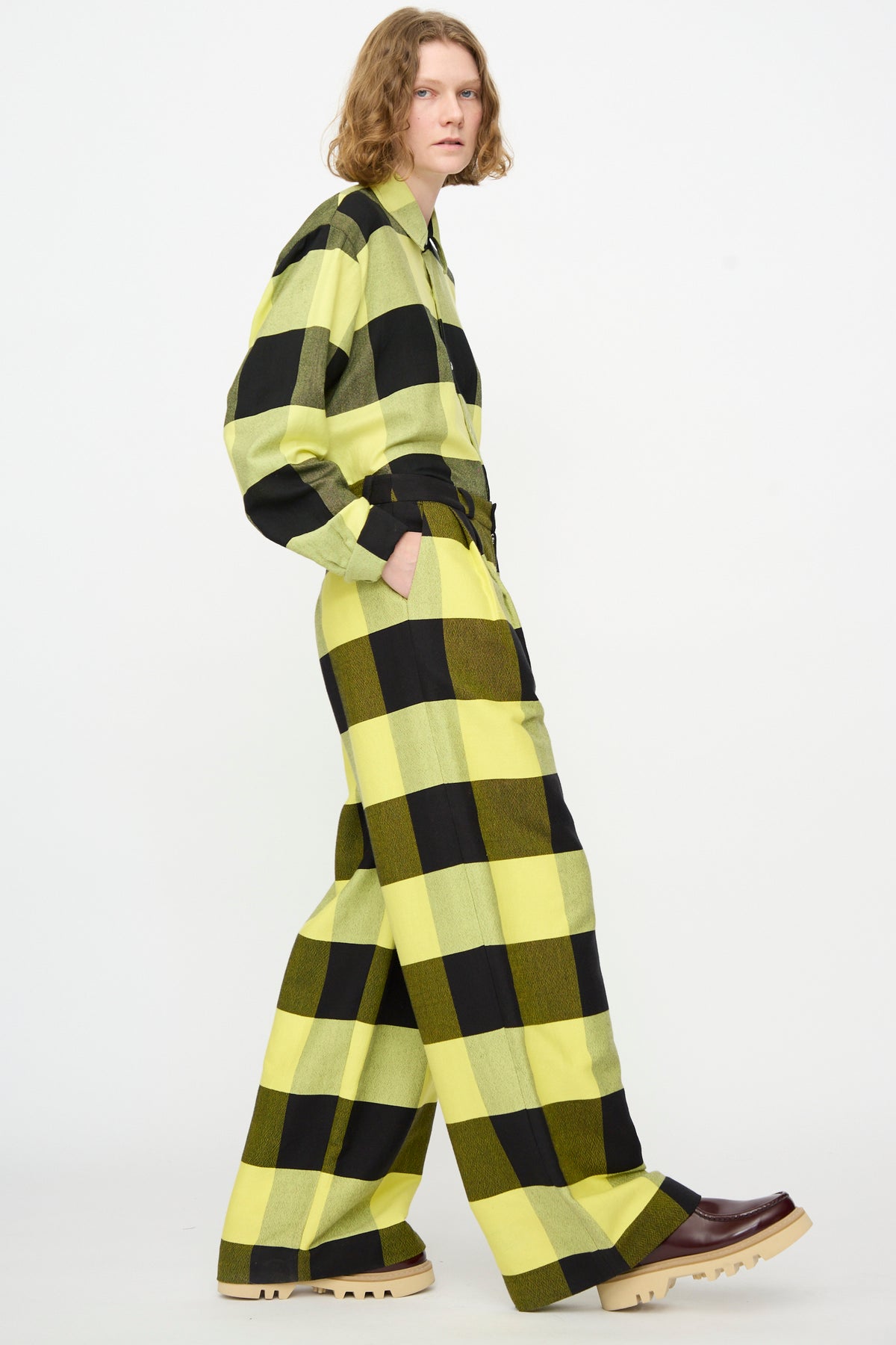 A person wearing Marrakshi Life’s “The Trouser in Plaid Black and Sunny”—a loose-fitting, wide-leg cotton plaid pant—styled with dark platform shoes, stands against a white background.