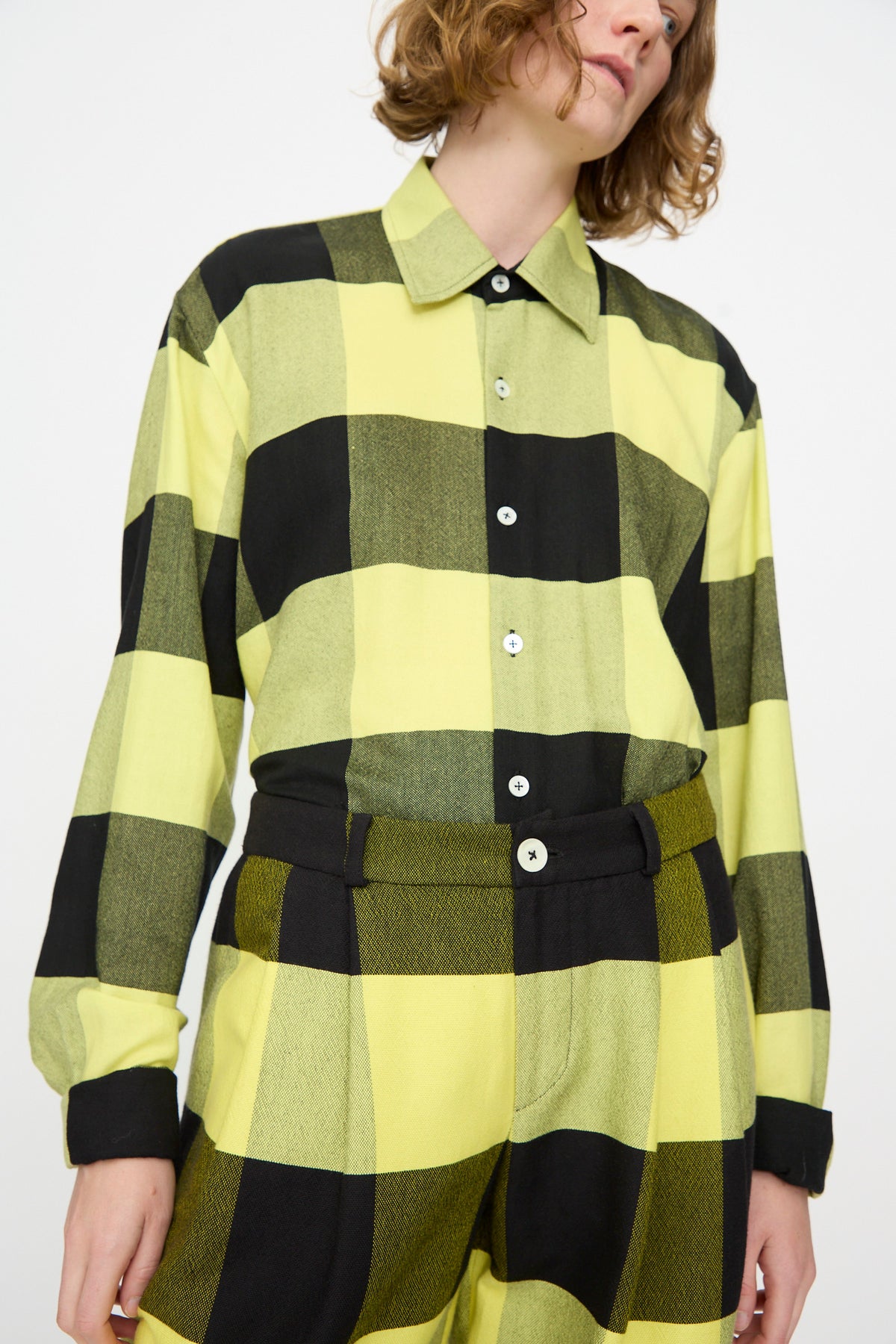 A person wearing Marrakshi Life’s The Trouser in Plaid Black and Sunny—a pair of high-waisted, plaid cotton trousers in black and yellow—tucked into a matching large-checkered shirt, standing against a plain white background.