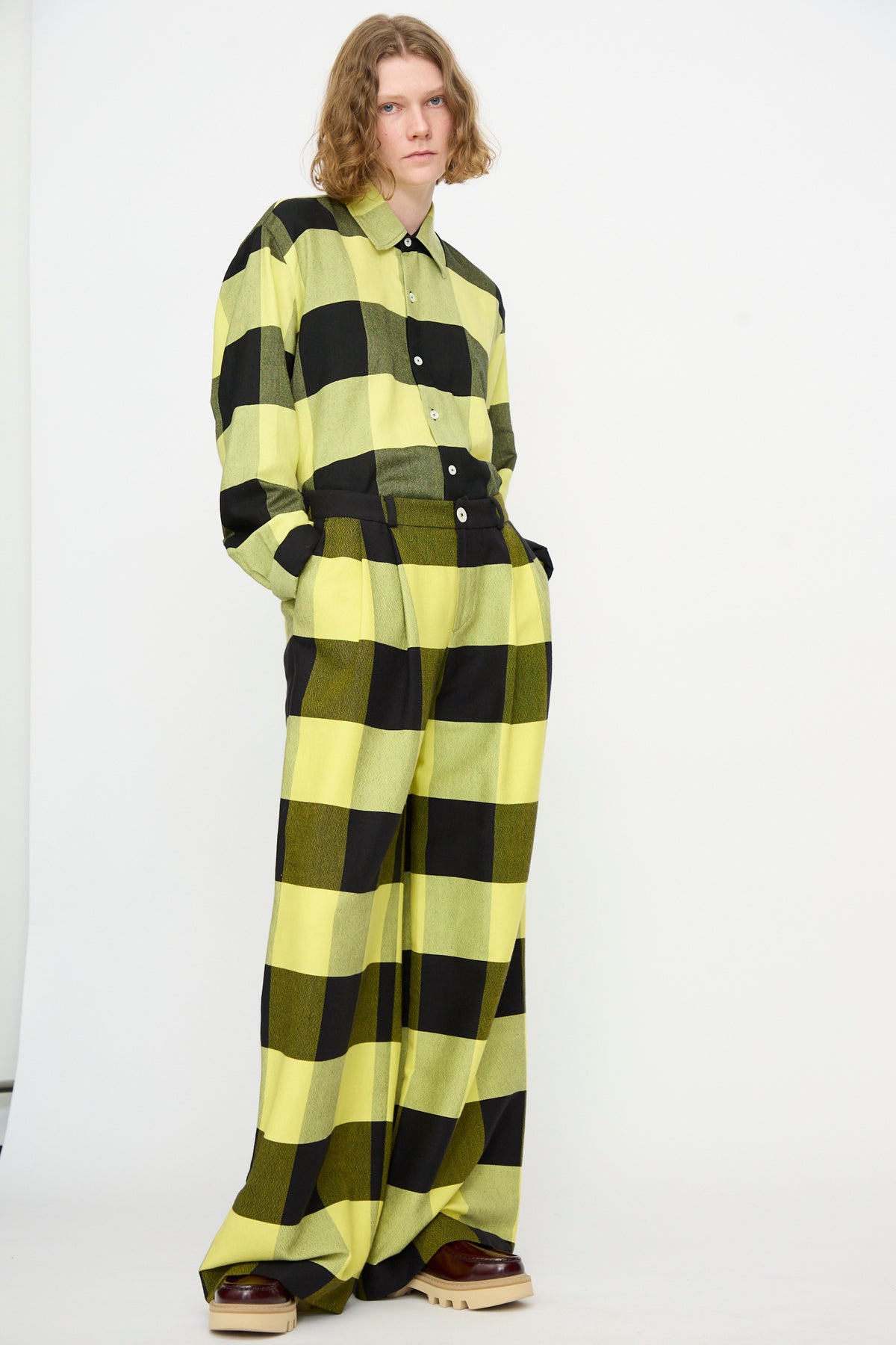 A person stands against a plain background wearing Marrakshi Life’s The Trouser in Plaid Black and Sunny—plaid cotton wide leg pants—paired with a matching yellow and black large checkered button-up shirt, hands in pockets, and brown platform shoes.