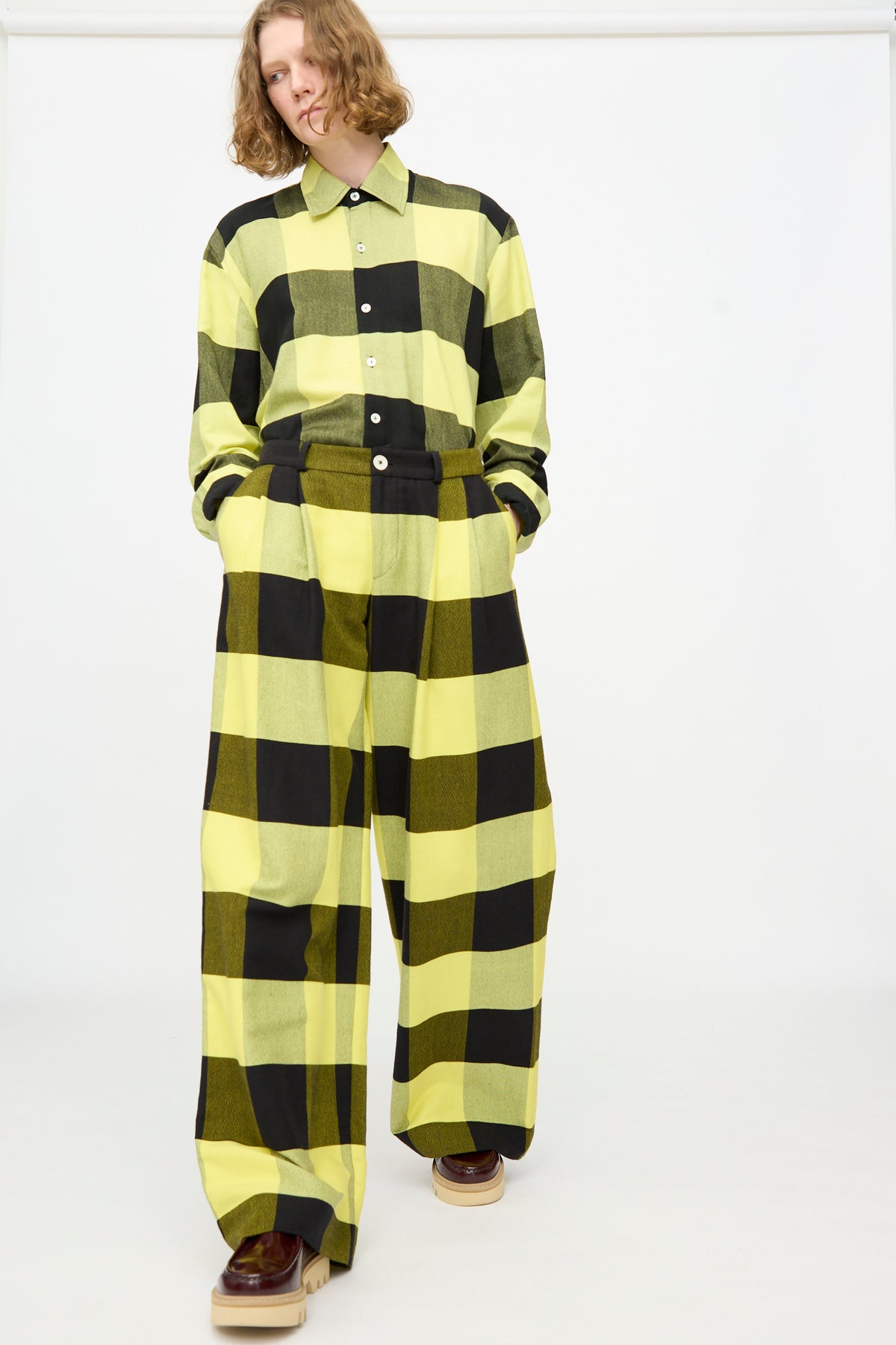 A person wearing The Trouser in Plaid Black and Sunny by Marrakshi Life—loose-fitting matching plaid cotton trousers paired with a yellow and black plaid shirt and brown platform shoes—stands against a plain white background.