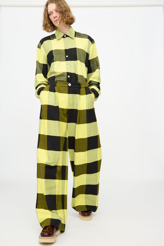 A person wearing The Trouser in Plaid Black and Sunny by Marrakshi Life—loose-fitting matching plaid cotton trousers paired with a yellow and black plaid shirt and brown platform shoes—stands against a plain white background.