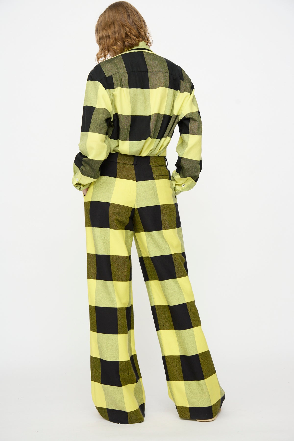 A person stands with their back to the camera, wearing Marrakshi Life’s The Trouser in Plaid Black and Sunny—wide leg cotton trousers featuring a yellow and black plaid pattern—paired with a matching checkered shirt, set against a plain white background.