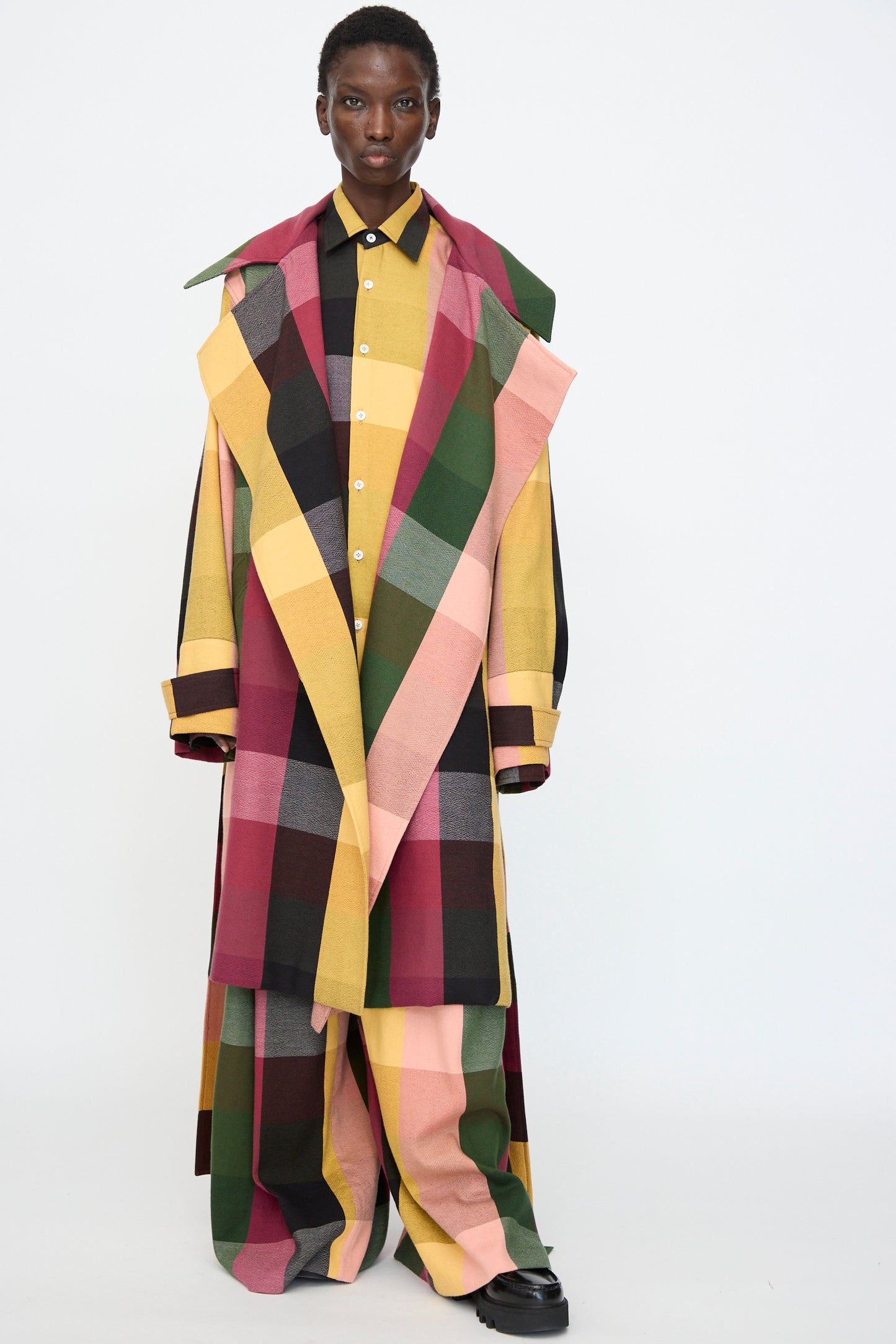 Model wears Marrakshi Life's Trench Coat in Plaid Multi—an oversized cotton piece in red, green, yellow, pink, and black—styled over a yellow button-up shirt and loose pants against a plain backdrop.