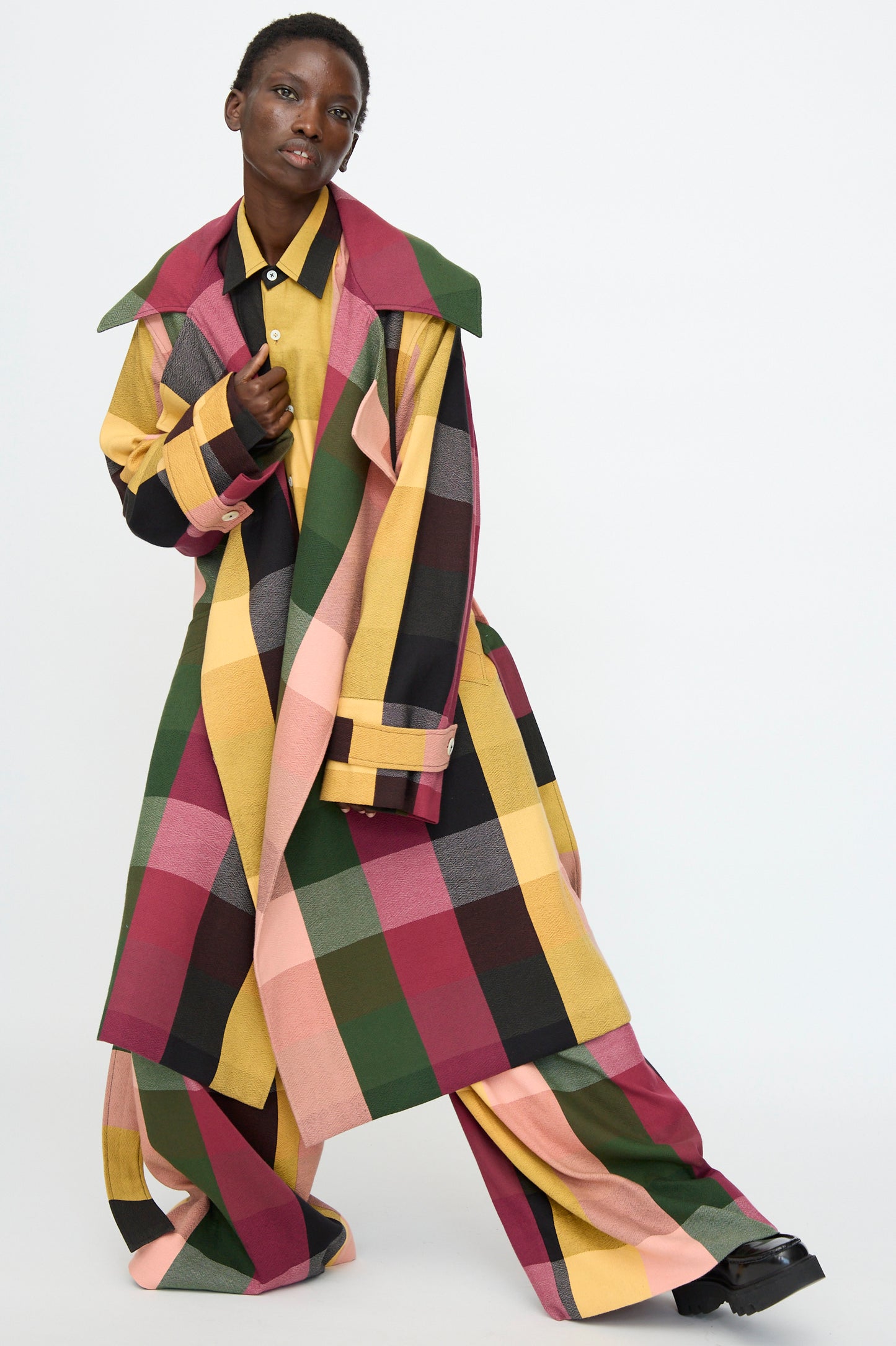 A person wears the Marrakshi Life Trench Coat in Plaid Multi with wide-leg pants, featuring vibrant yellow, pink, green, black, and burgundy plaid, standing against a plain white background.