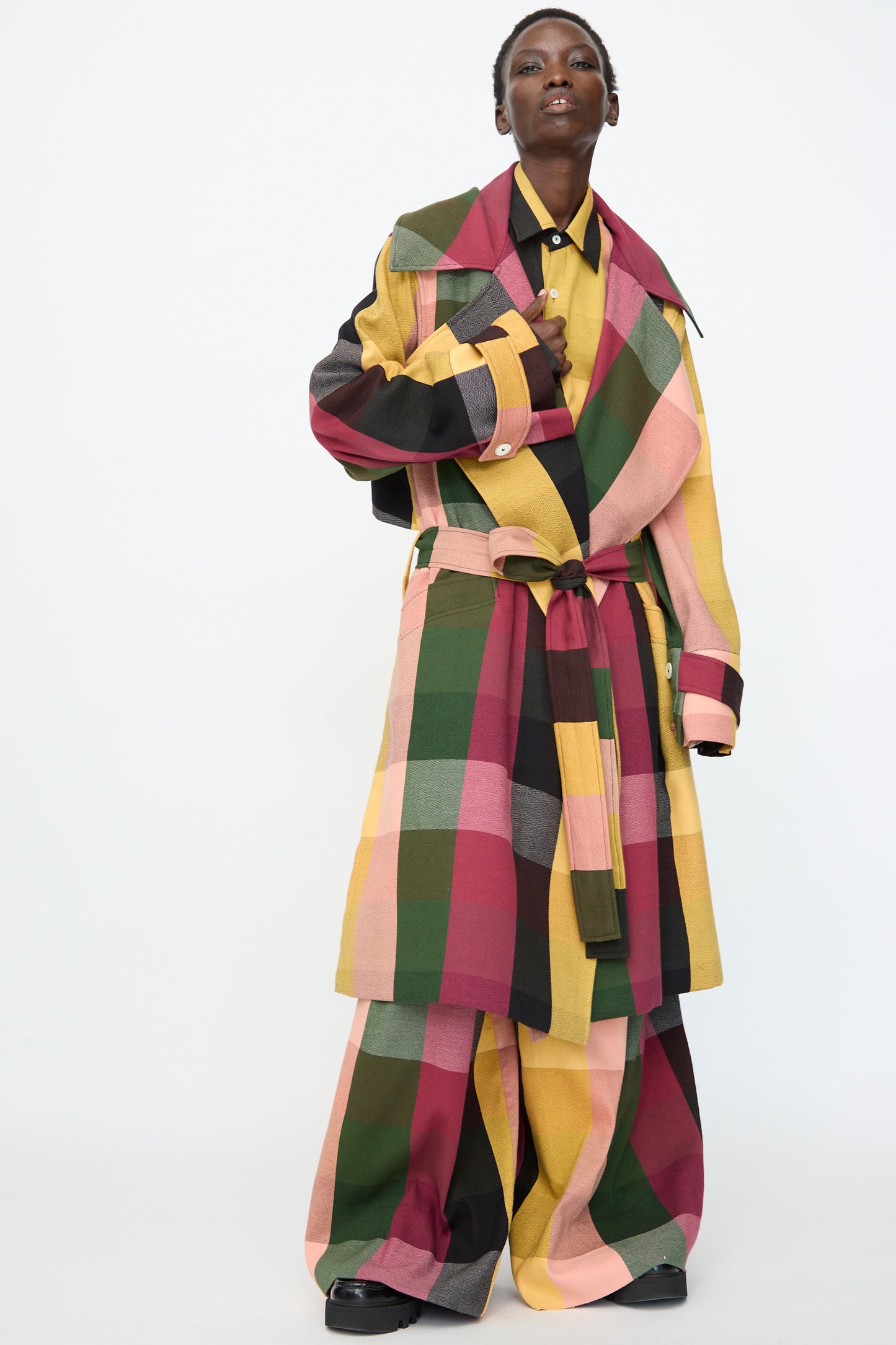 A person models the Marrakshi Life Trench Coat in Plaid Multi with matching wide-leg pants in green, yellow, pink, and maroon tones, standing against a plain white background.