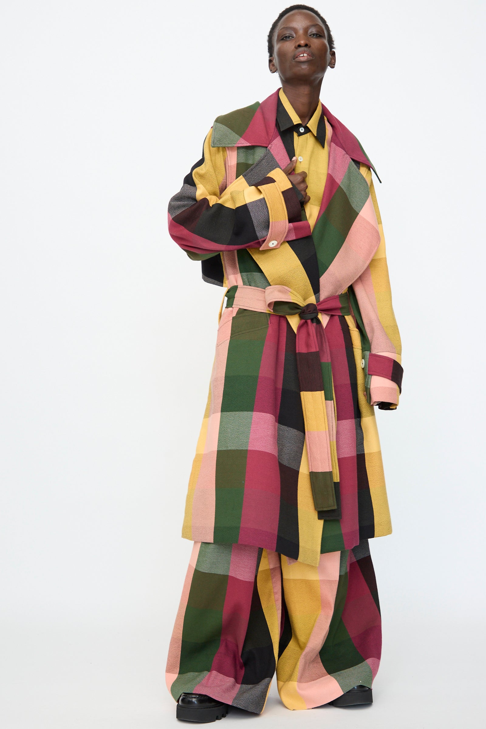 A person models the Marrakshi Life Trench Coat in Plaid Multi with matching wide-leg pants in green, yellow, pink, and maroon tones, standing against a plain white background.