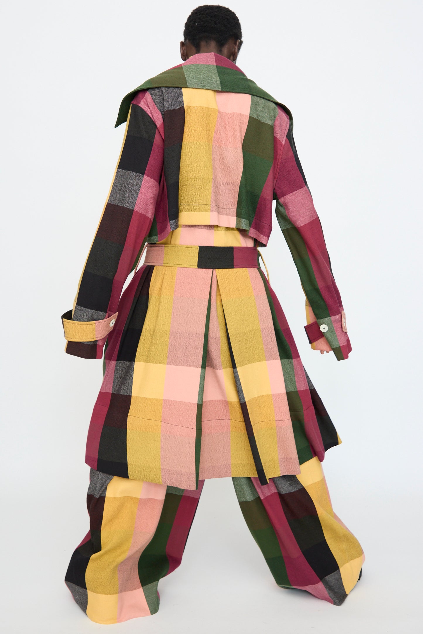 A person stands with their back turned, wearing the Marrakshi Life Trench Coat in Plaid Multi— a cotton trench coat featuring wide pants and pleated details in pink, yellow, black, and green.