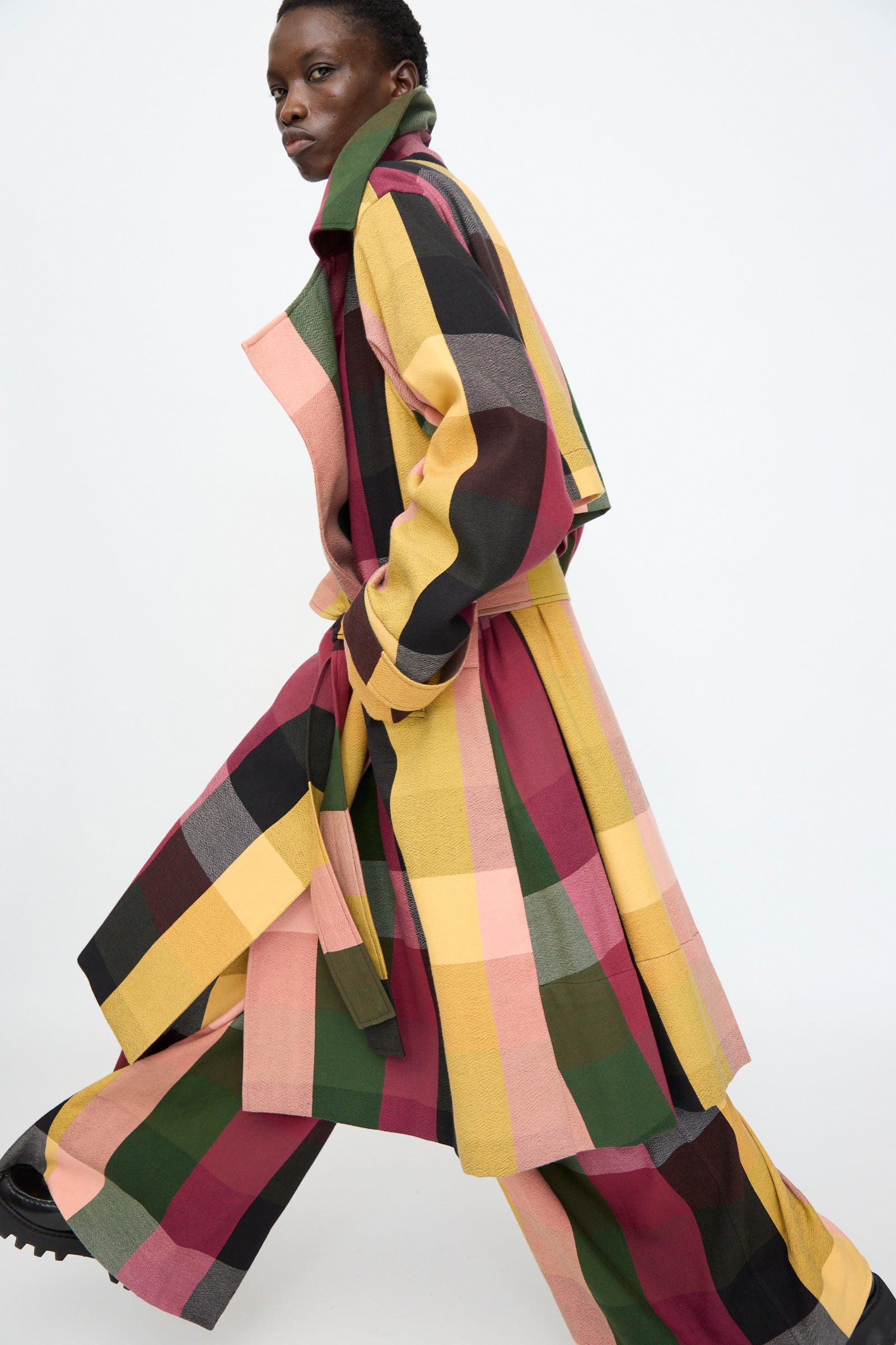 A person wearing Marrakshi Life’s Trench Coat in Plaid Multi walks forward against a plain white background.