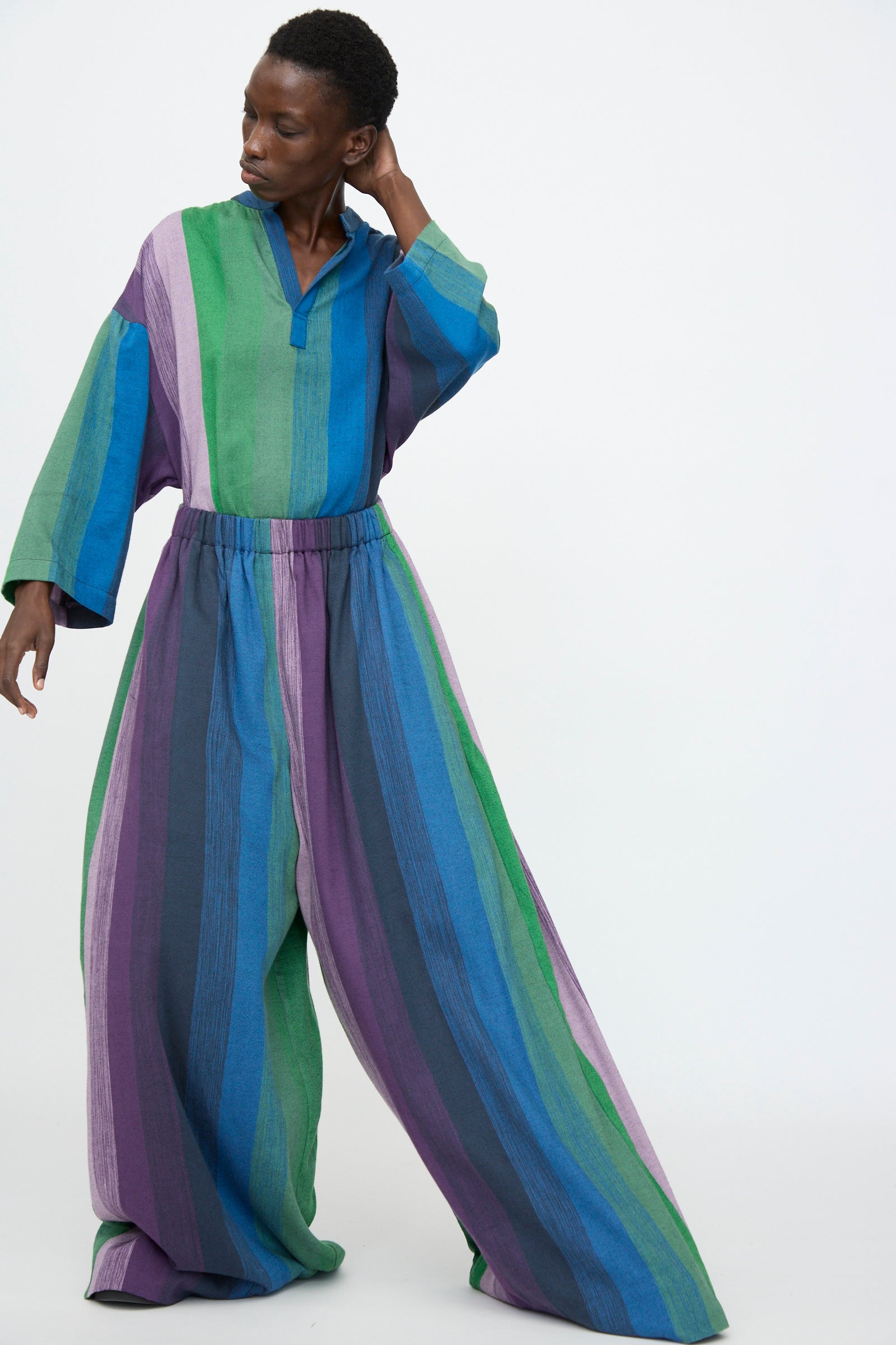 A person wears the Marrakshi Life Wide Leg Pant in Stripe Multi—loose, striped cotton pants with an elastic waist and oversized legs in green, blue, purple, and gray—standing against a plain white background.