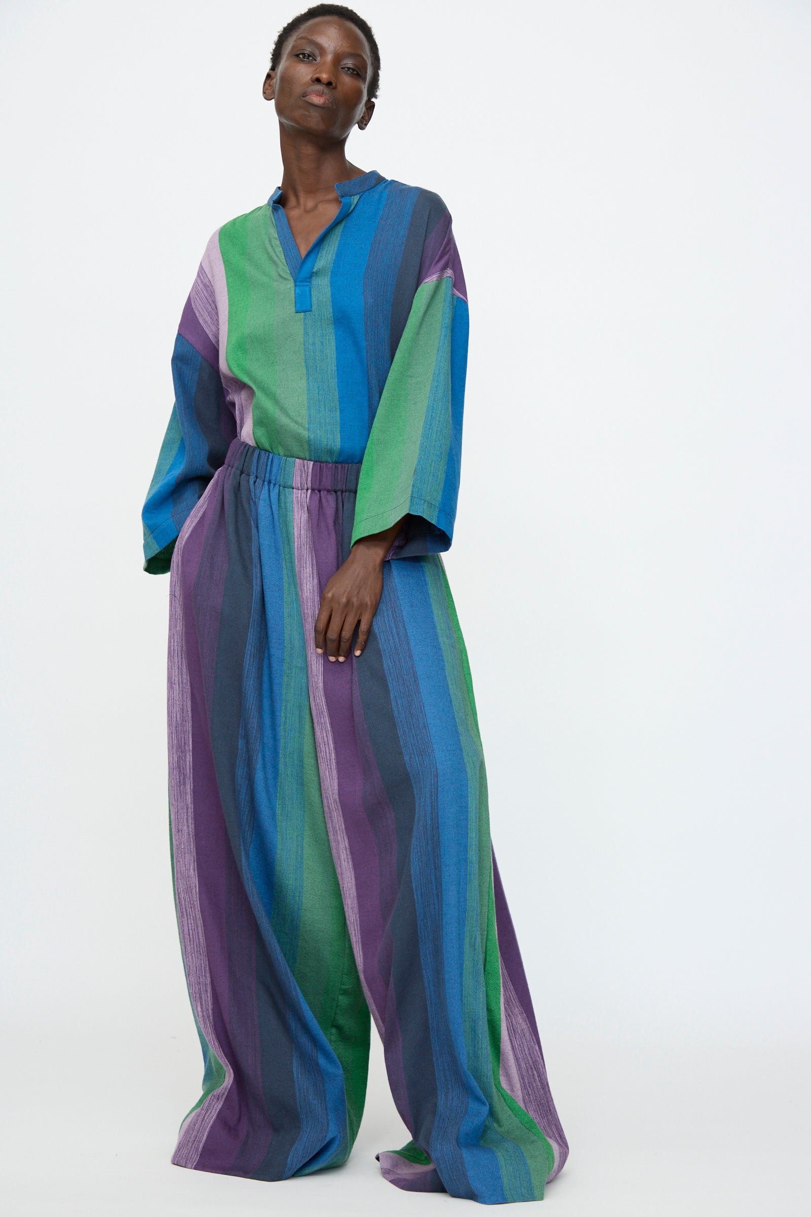 A person wearing Marrakshi Life's Wide Leg Pant in Stripe Multi, featuring vertical blue, green, and purple stripes, stands against a plain white background.