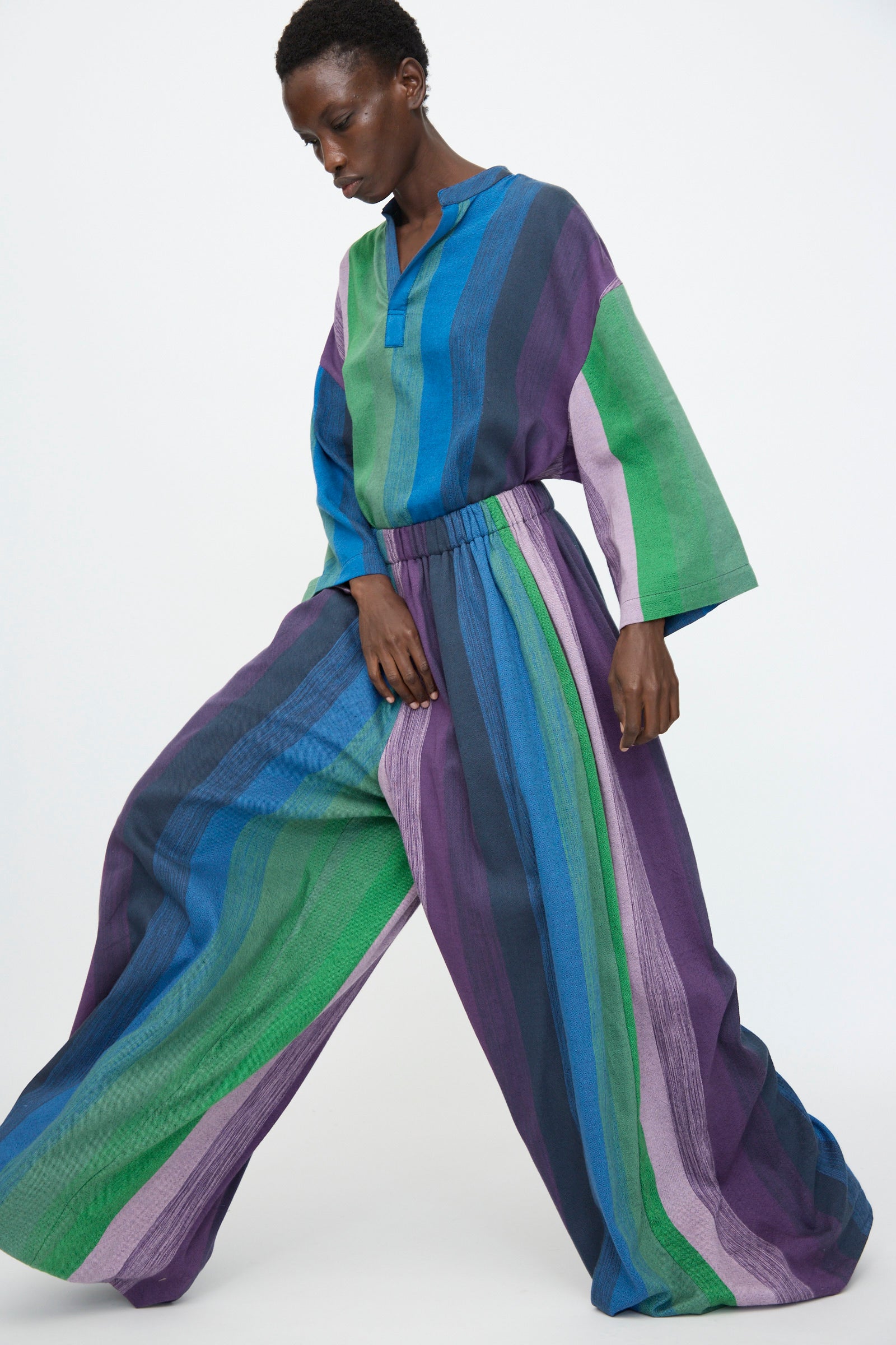 A person stands against a white background wearing Marrakshi Life's Wide Leg Pant in Stripe Multi—an oversized, elastic-waist cotton pant in blue, purple, green, and teal—with a loose-fitting long-sleeved top.