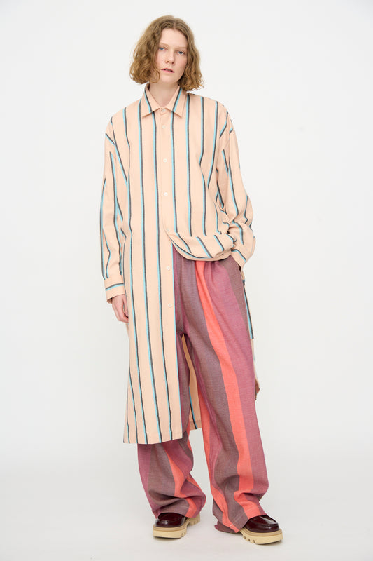 A person wears Marrakshi Life’s The Long Shirt in Stripe—a long, beige handwoven cotton shirt with vertical stripes—paired with loose, wide-legged pink pants featuring vertical red stripes, standing against a plain white background. This oversized long sleeve shirt is handmade in Morocco.