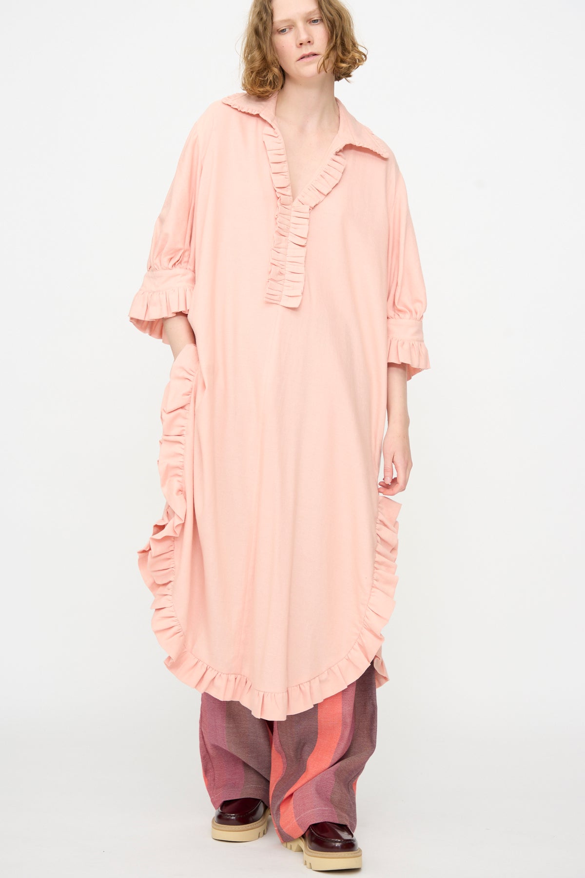A person wears The Ruffled Tunic in Flesh by Marrakshi Life—an oversized, light pink, handmade dress from Morocco—paired with wide-legged striped pants and platform shoes, standing against a plain white background.