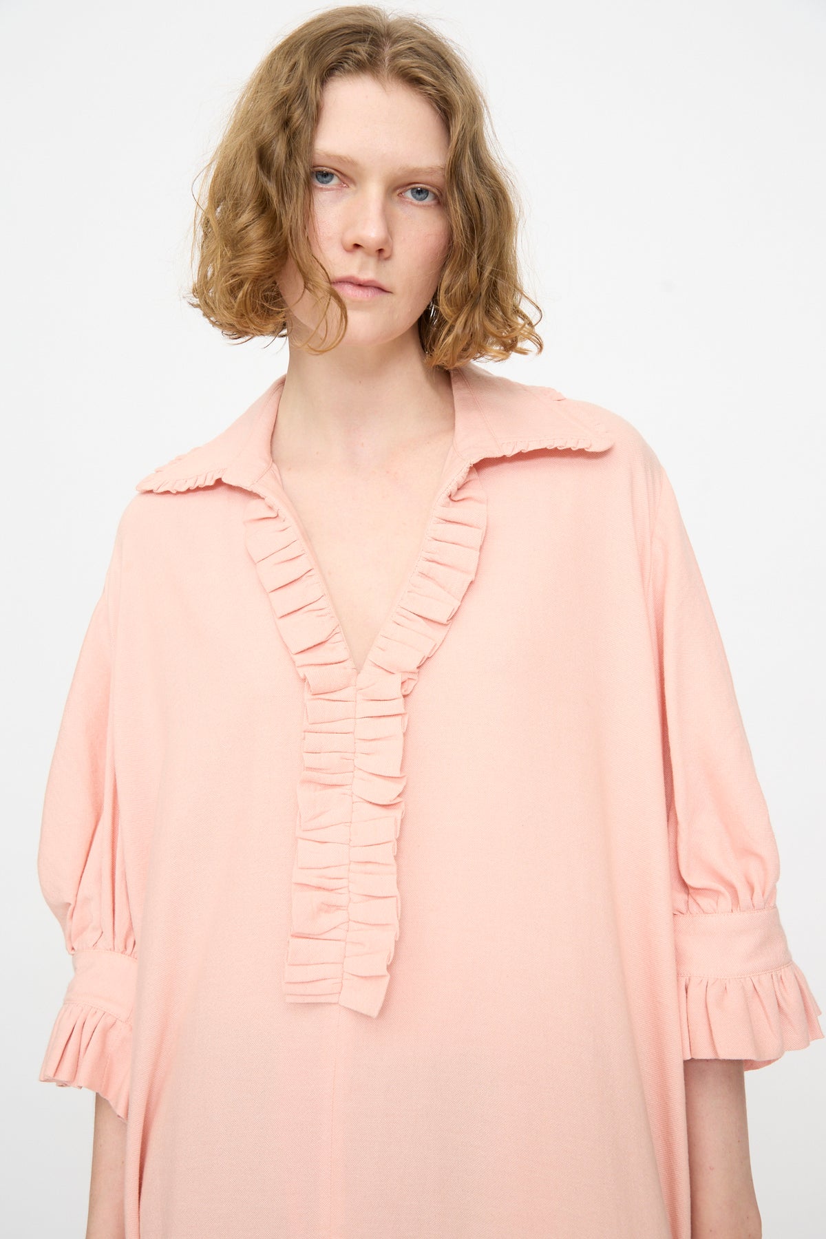 A person with short, wavy blond hair wears Marrakshi Life’s The Ruffled Tunic in Flesh—a loose, light pink handwoven cotton tunic with ruffled trim along the collar, sleeves, and front—standing against a plain white background.