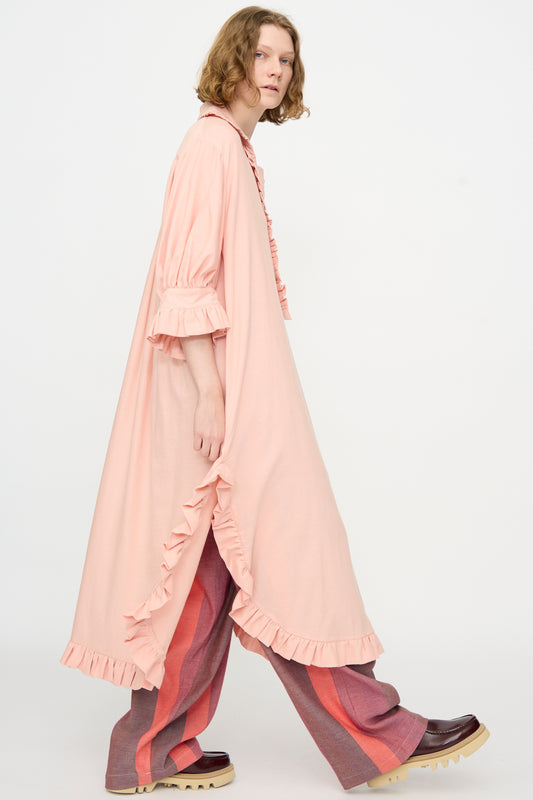 A person is wearing Marrakshi Life’s The Ruffled Tunic in Flesh, styled as an oversized light pink dress over wide-leg pants featuring bold pink and red stripes, paired with chunky brown shoes, walking against a plain white background.