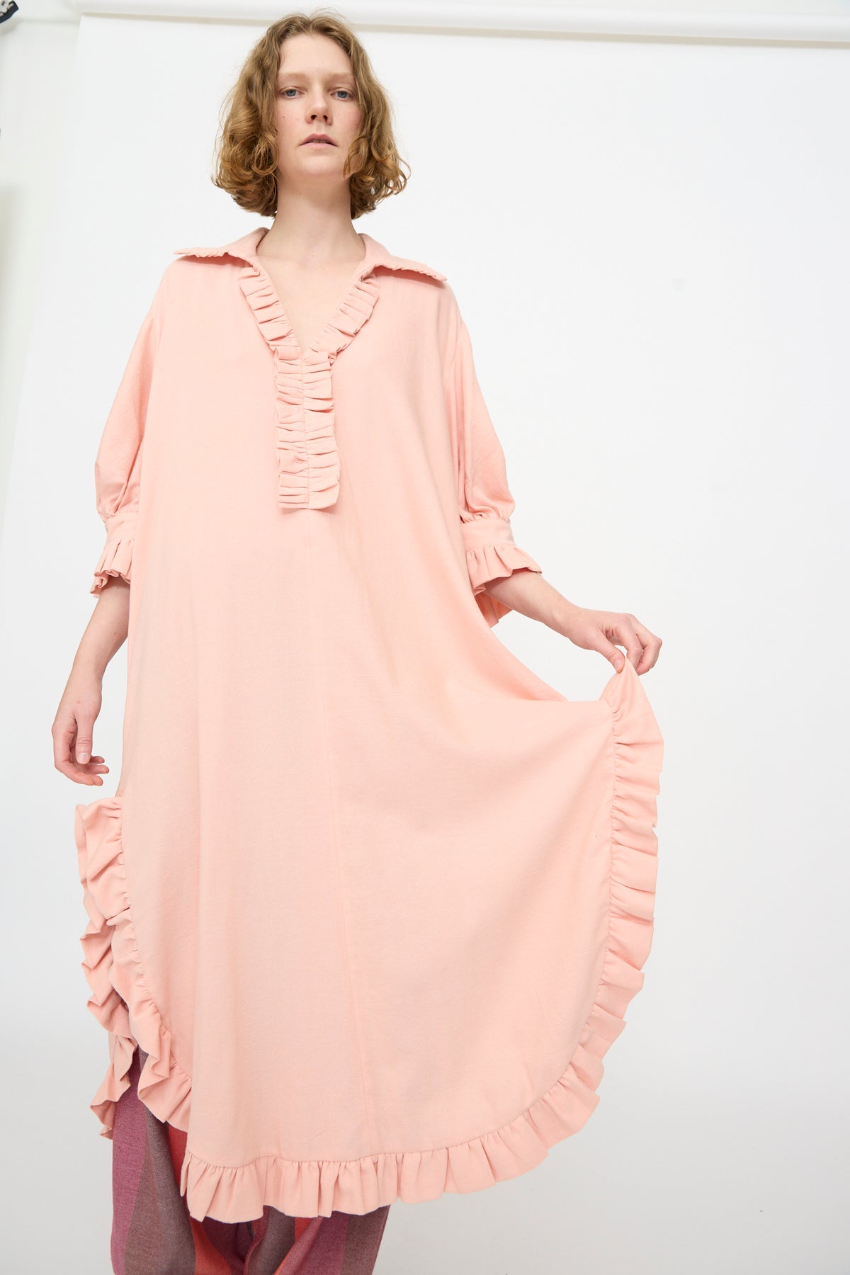 A person is wearing Marrakshi Life’s The Ruffled Tunic in Flesh—an oversized, light pink dress crafted with ruffled edges and long sleeves—standing against a white background.