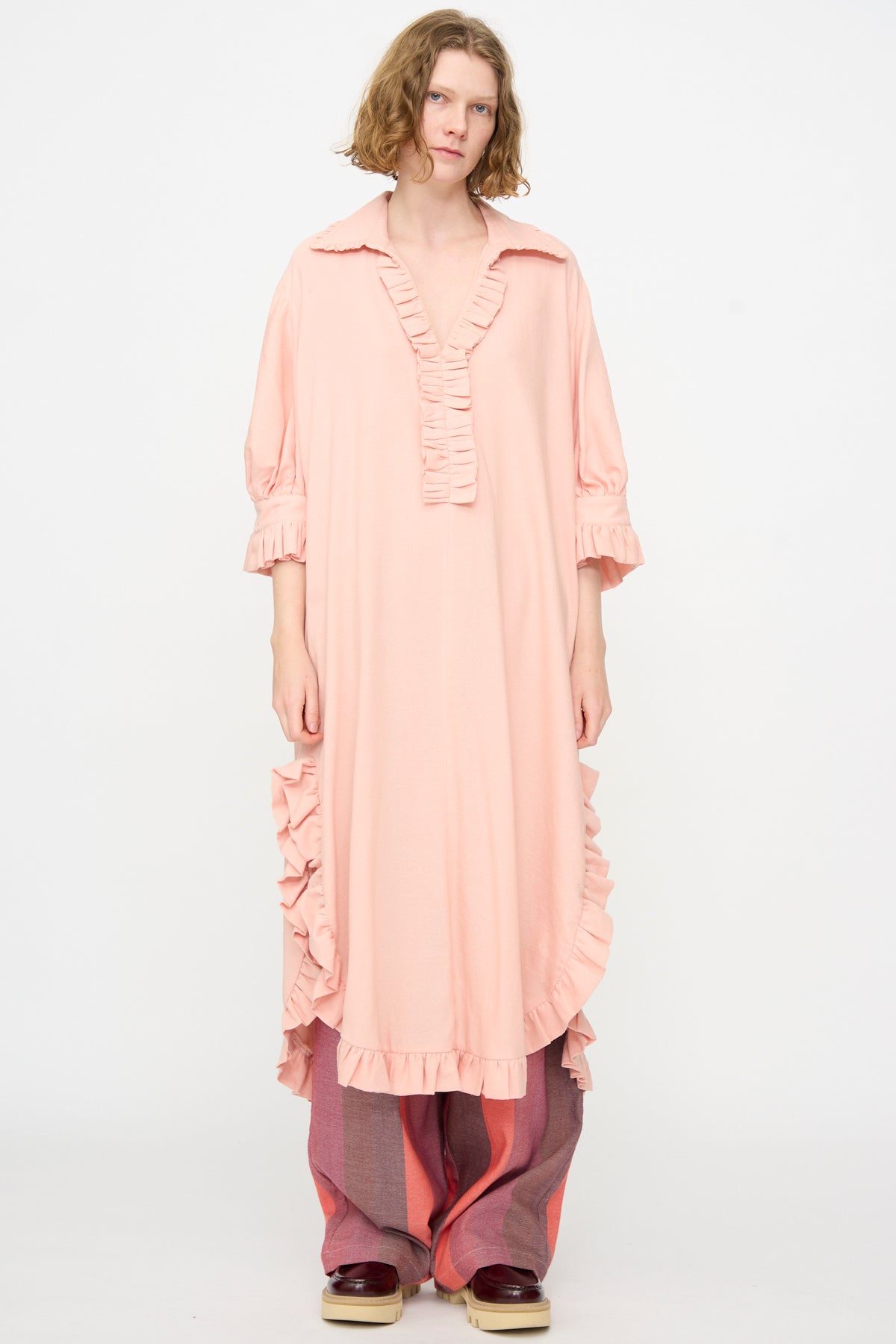 A person wears Marrakshi Life’s The Ruffled Tunic in Flesh—a loose, light pink ruffled dress—layered over wide-legged pink and red pants, standing against a plain white background.