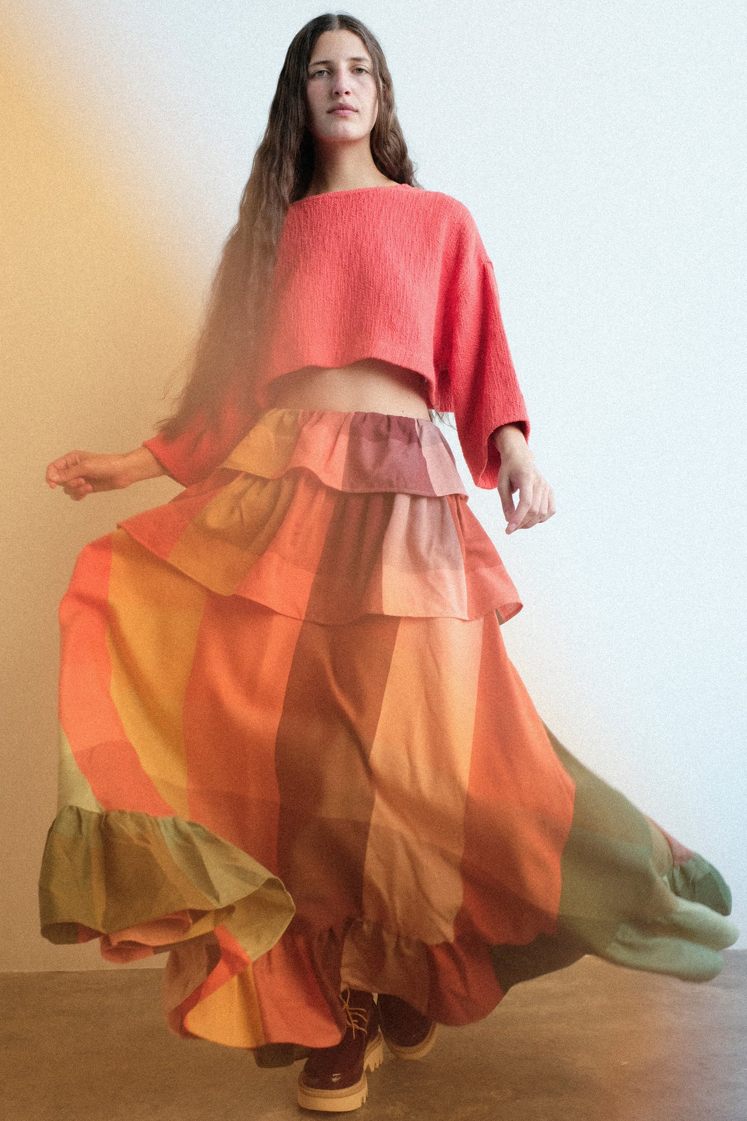A woman wearing Marrakshi Life's Cropped T-Shirt in Burnt Orange Flamme, and The Tiered Ruffle Skirt in Plaid, captured in motion against a plain light background, with an orange filter on the left.