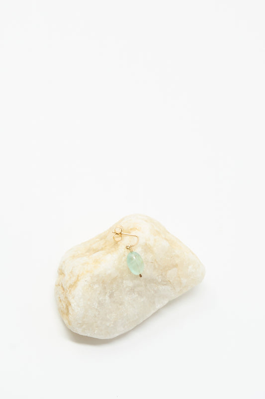 The Mary MacGill 14K Crescent Charm Single Earring in Aquamarine Stone is displayed on a white stone, with its gold hoop and translucent blue-green aquamarine bead set against a plain white background.