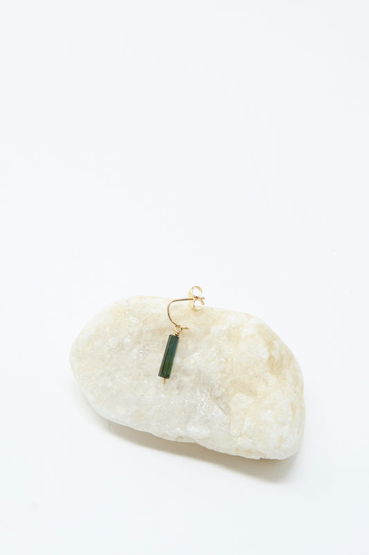 The Mary MacGill 14K Crescent Charm Single Earring in Dark Tourmaline Tube, featuring a handmade vertical green stone, is displayed on a smooth light-colored rock against a white background.