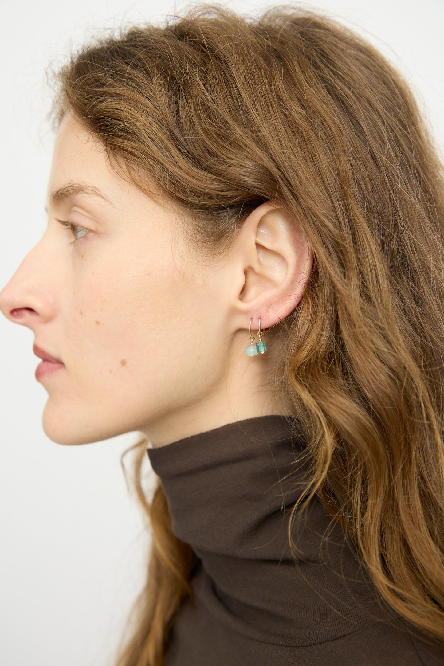 A woman with wavy light brown hair in a dark turtleneck faces left in profile, wearing Mary MacGill's 14K Crescent Charm Single Earring featuring a faceted green emerald, set against a plain background.
