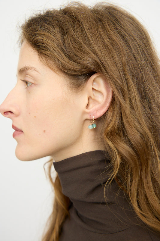 A woman with wavy light brown hair in a dark turtleneck faces left in profile, wearing Mary MacGill's 14K Crescent Charm Single Earring featuring a faceted green emerald, set against a plain background.