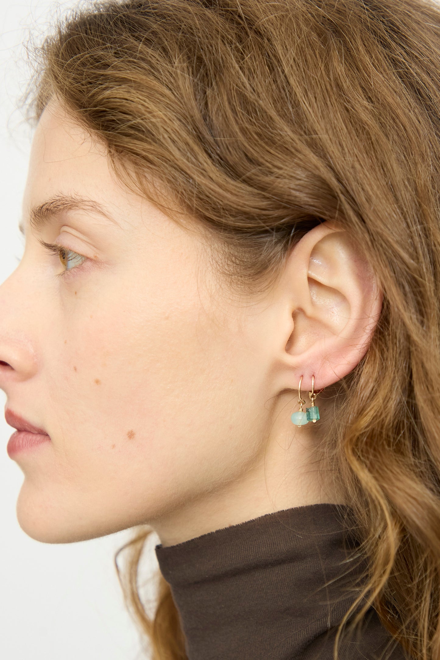 A woman with wavy brown hair wears the Mary MacGill 14K Crescent Charm Single Earring in Faceted Green Emerald, shown in profile against a plain background.