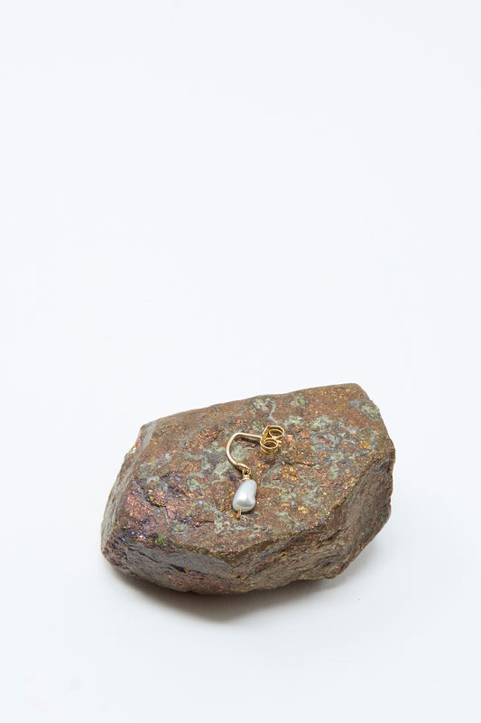 The Mary MacGill 14K Crescent Charm Single Earring in Keshi Pearl rests atop a textured brown rock, showcased against a plain white background.