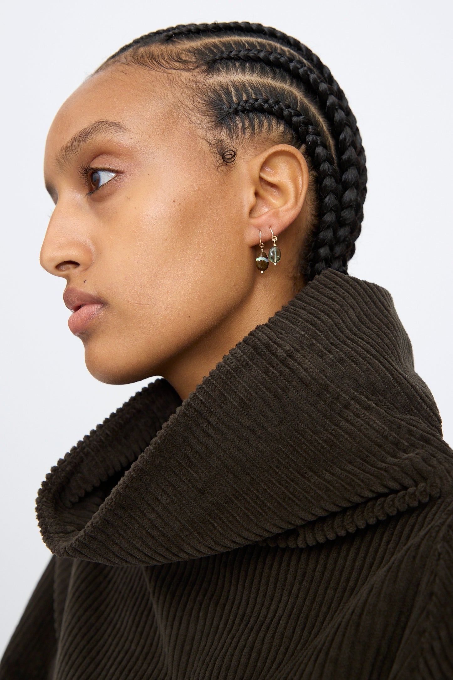 A person with cornrow braids in a dark ribbed turtleneck wears the Mary MacGill 14K Crescent Charm Single Earring in Boulder Opal, shown in profile against a plain background.