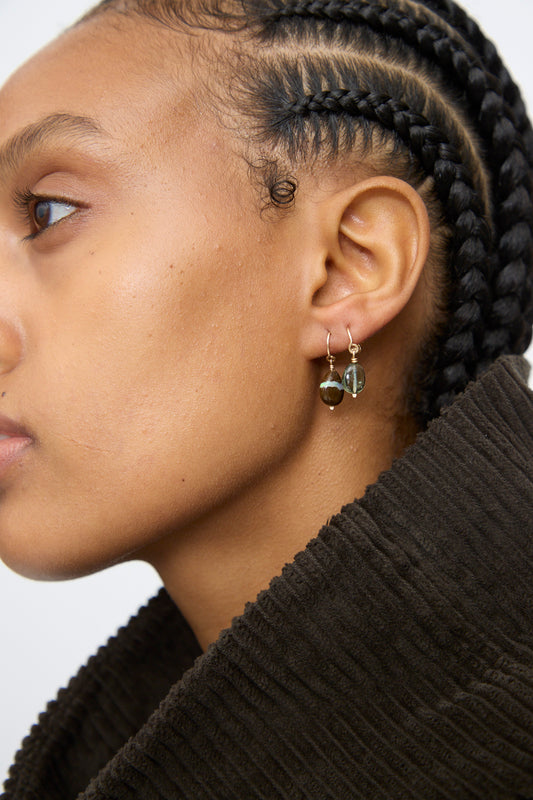 Close-up of a person with braided hair in a dark textured jacket, wearing Mary MacGill's 14K Crescent Charm Single Earring in Boulder Opal, facing left.