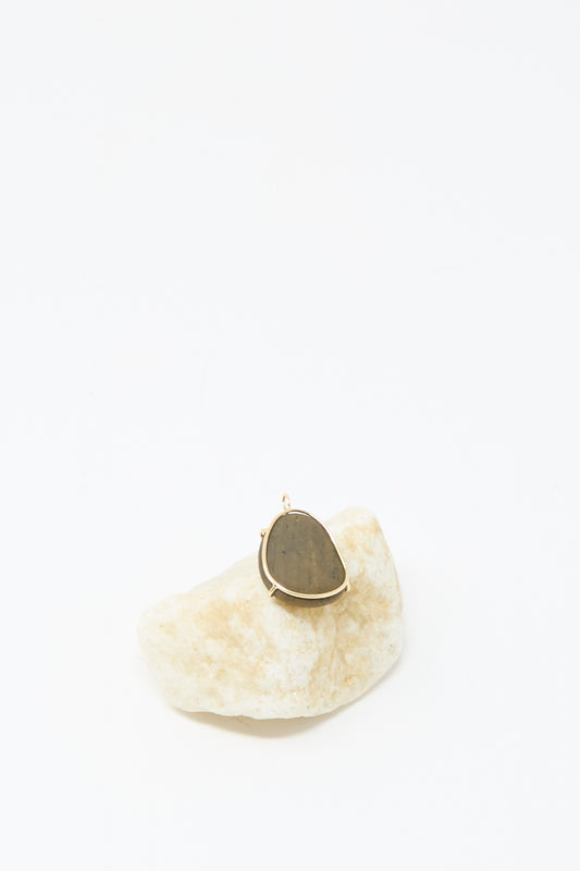 The Mary MacGill 14K Floating Pendant in Island Stone, featuring a handmade teardrop dark stone set in 14k gold, is displayed on a light rock against a white background.