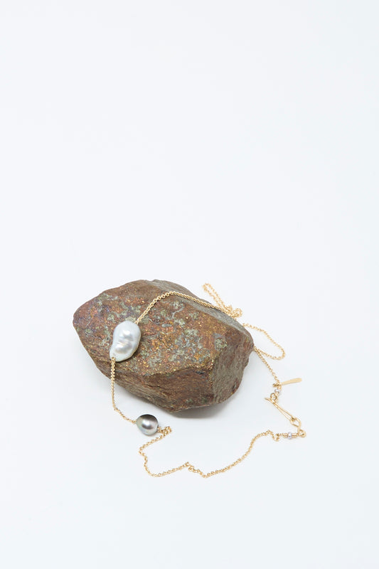 The Mary MacGill 14K Necklace featuring White Keshi and Silver Tahitian Pearls is draped over a rough brown stone against a plain white background.