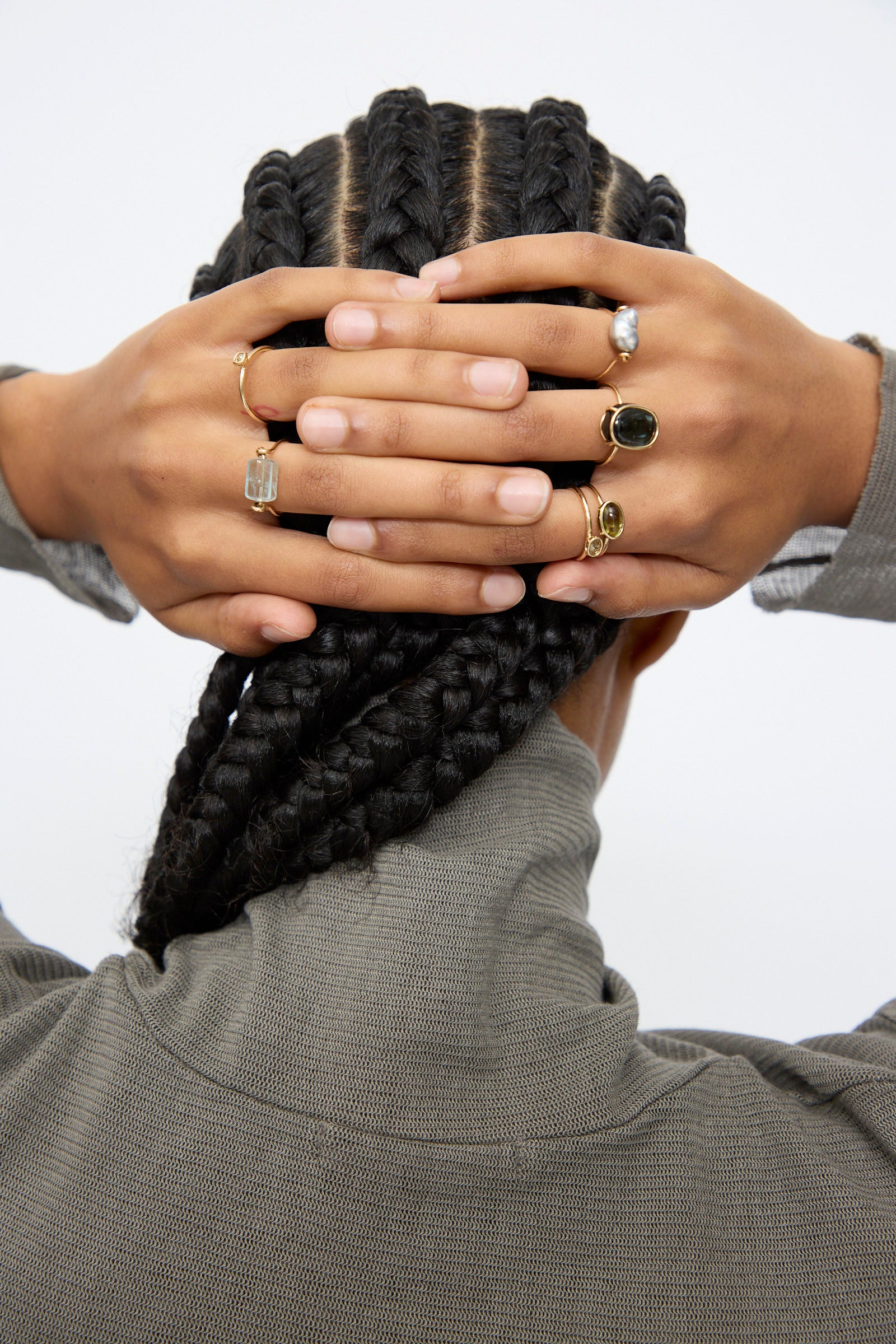 A person with cornrow braids seen from behind, hands on the back of their head, wearing multiple handmade gold rings—including the Mary MacGill 14K Ring in Aquamarine Tube—and a gray textured top.