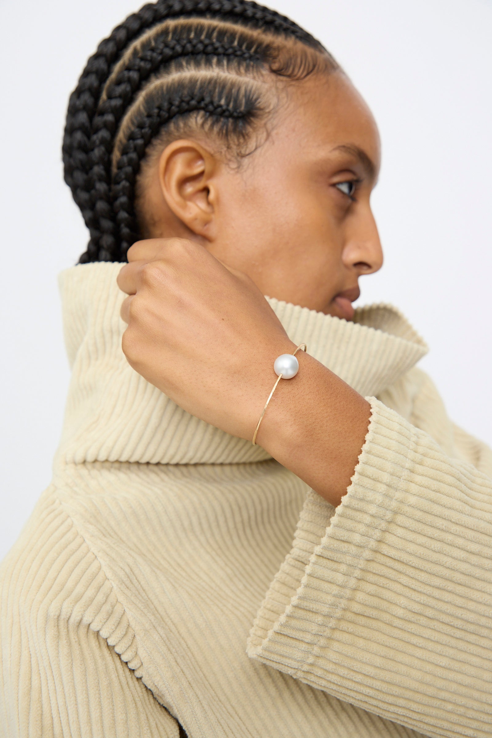 A person with cornrow braids wears a beige ribbed turtleneck sweater and the Mary MacGill 14K Tahitian Pearl Cuff in White.