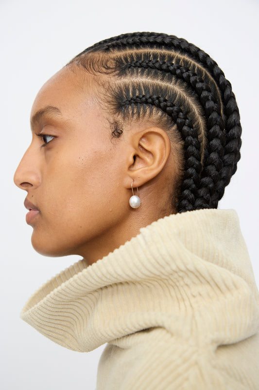 Profile view of a person with cornrow braids, wearing a cream textured turtleneck and Mary MacGill's 14K Tahitian Pearl Drop Earrings in White.