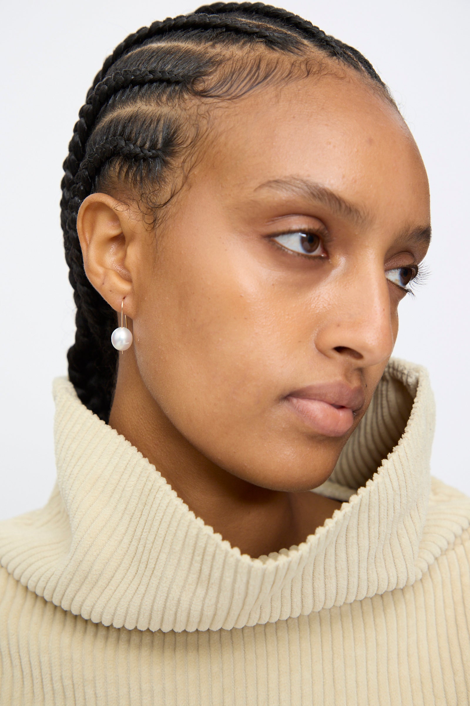 A person with cornrow braids, wearing a beige ribbed turtleneck and a single Mary MacGill 14K Tahitian Pearl Drop Earring in White, looks slightly to the side.