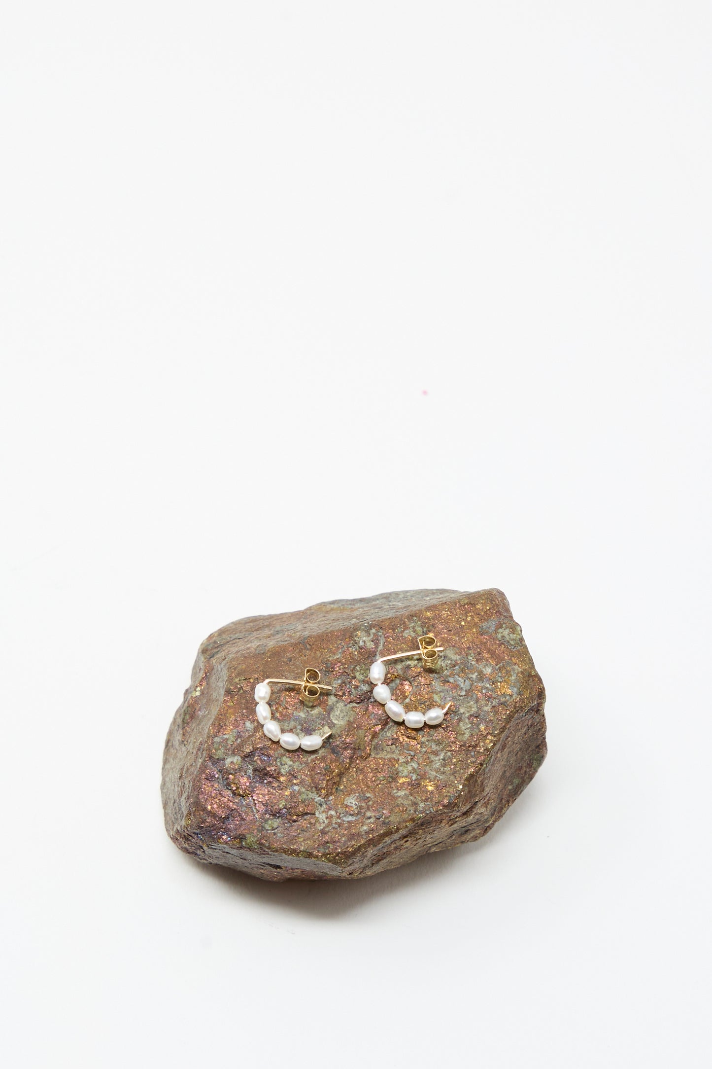 The Mary MacGill Baroque Pearl Crescent Hoops, featuring handmade baroque pearls and gold hoops, are displayed on a rough textured stone with a plain white background.