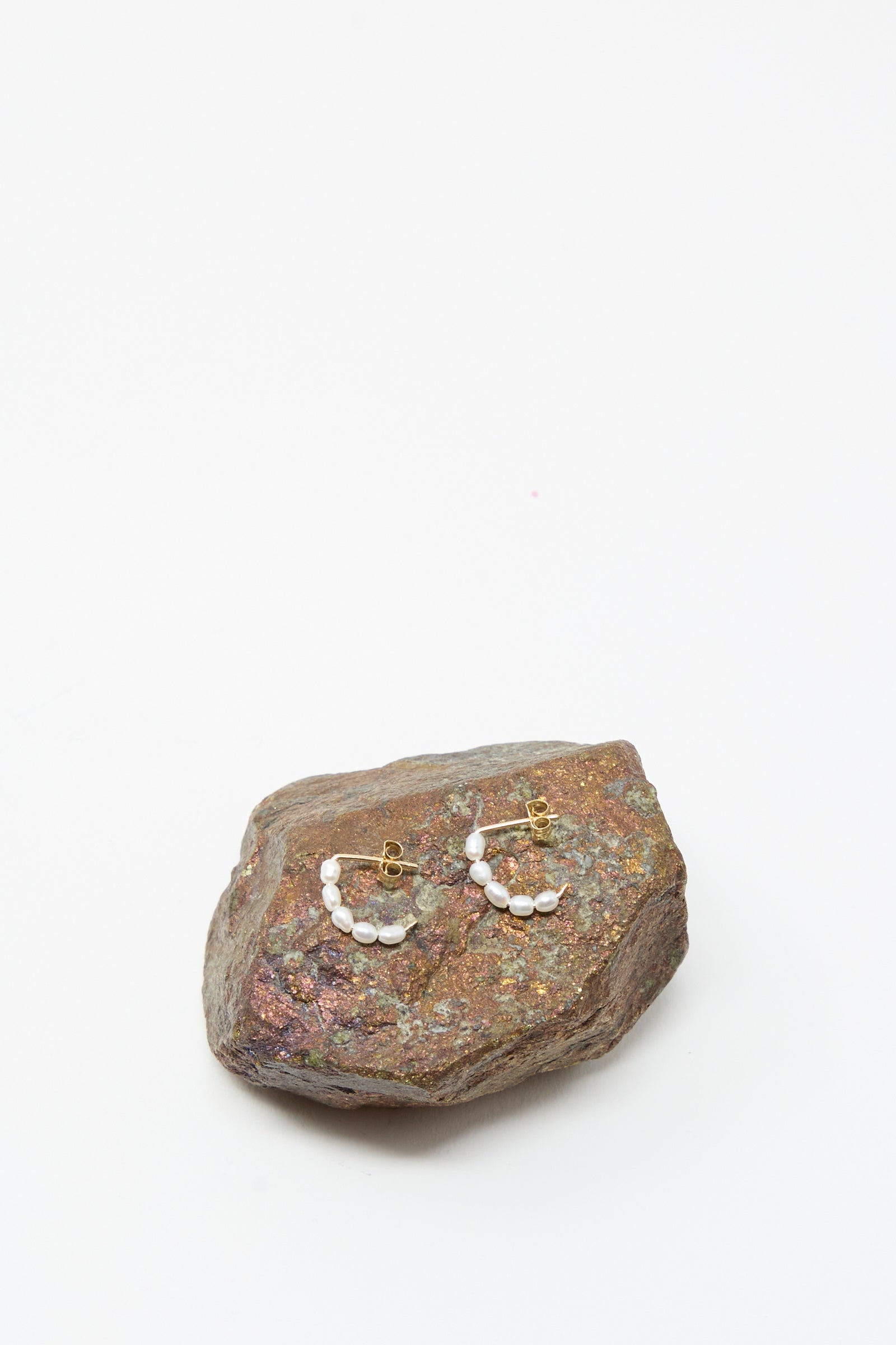 The Mary MacGill Baroque Pearl Crescent Hoops, featuring handmade baroque pearls and gold hoops, are displayed on a rough textured stone with a plain white background.