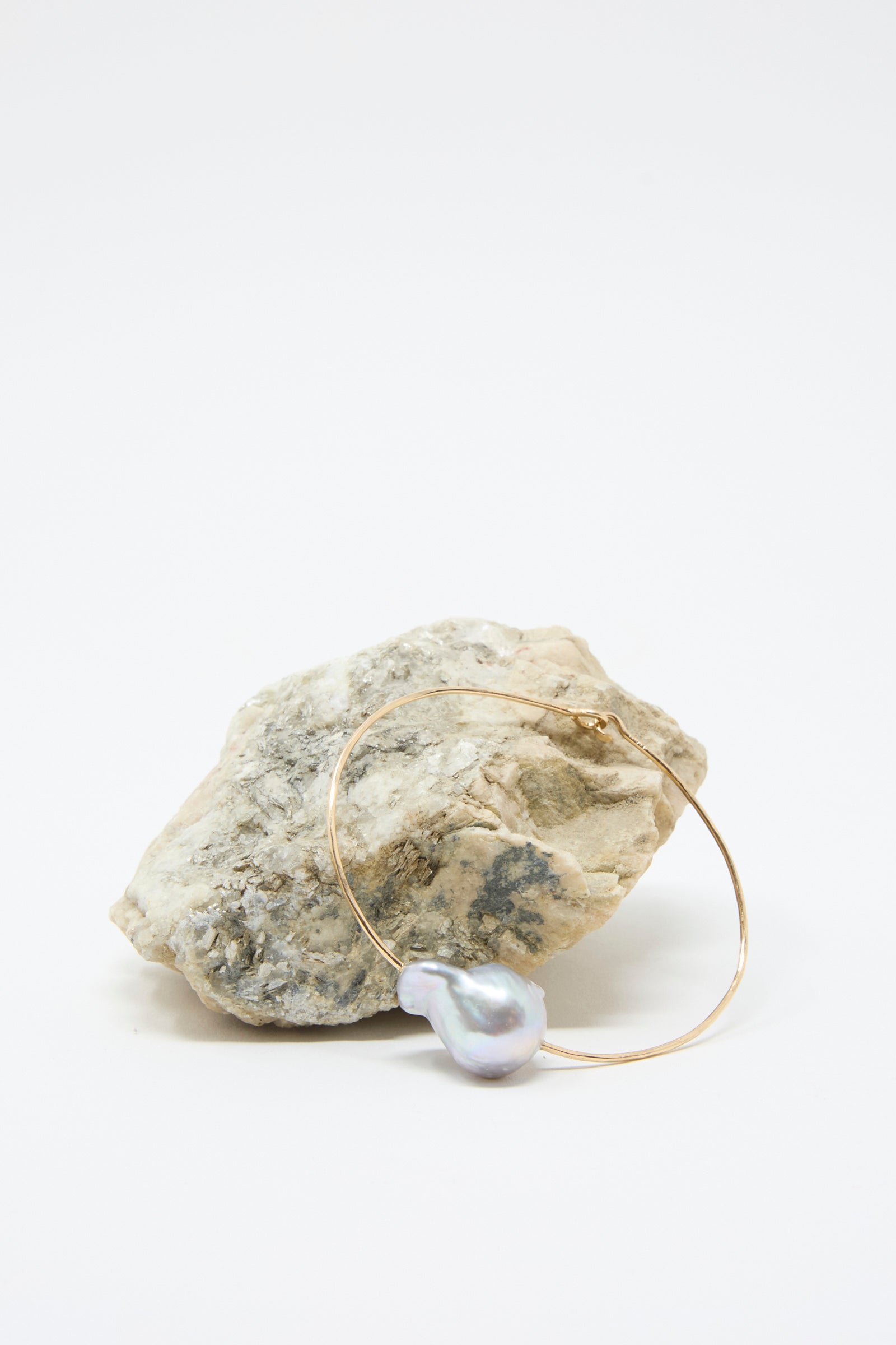 A Baroque Pearl Cuff Bracelet in Silver by Mary MacGill is positioned leaning against a rough-textured stone against a white background. 